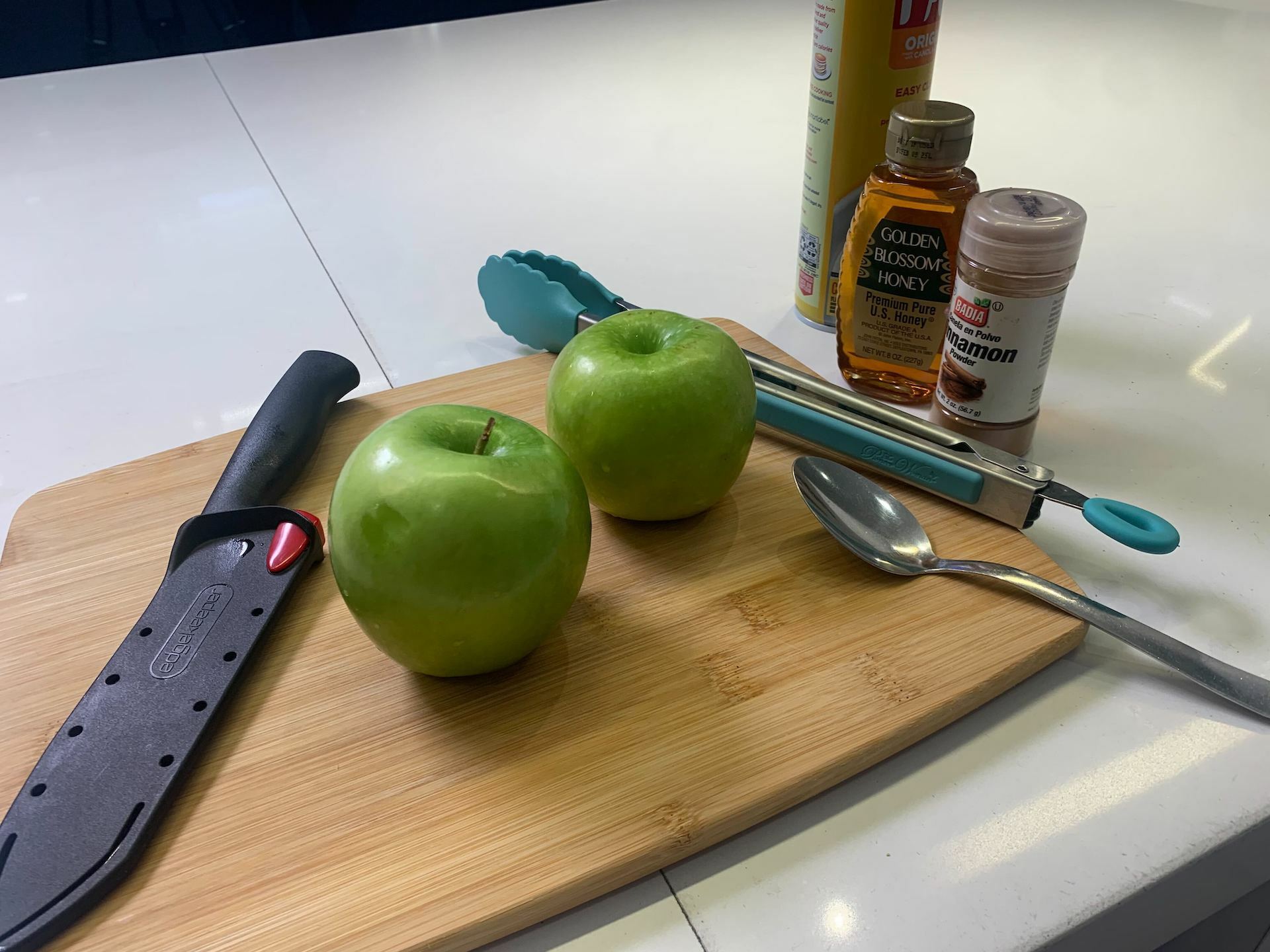 a knife, apples, tongs, spoon, honey, and cinnamon on a cutting baord