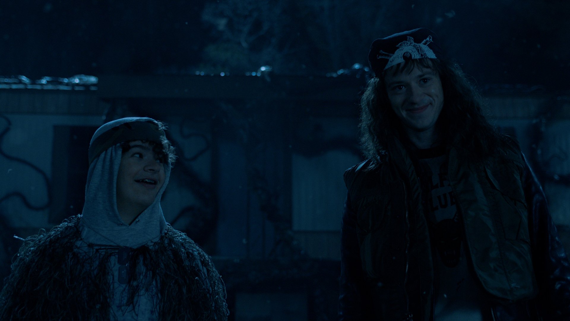 Two teens stand in a grim, blue-lit world but still smiling.