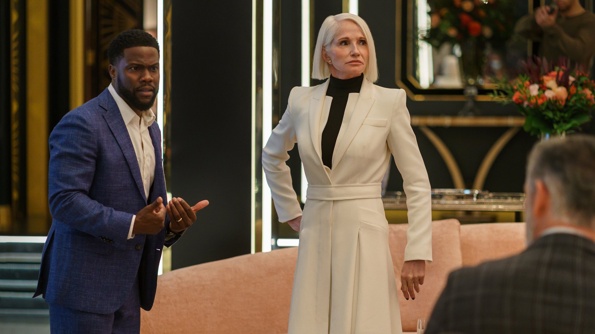 Two people in suits stand looking worried in a posh living room.