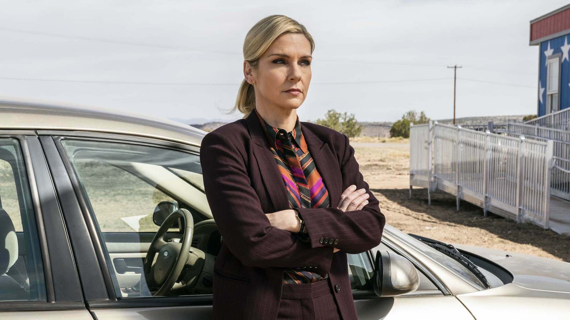 A woman in a suit leans against a car next to a building in the desert.