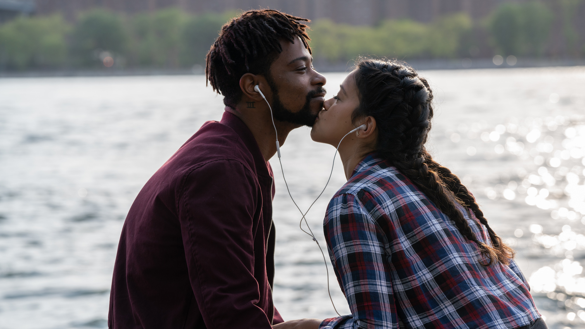 Two people sharing an earphone each sit by a river. One kisses the other on the chin.