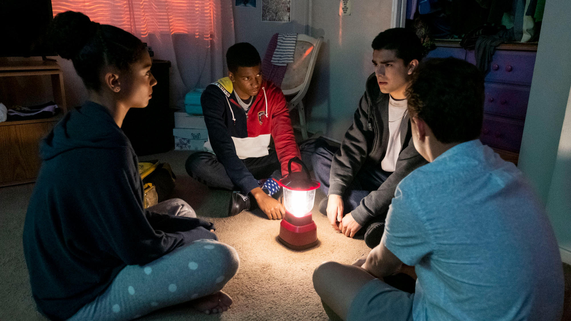 A group of teens sit in a bedroom around a camp light.
