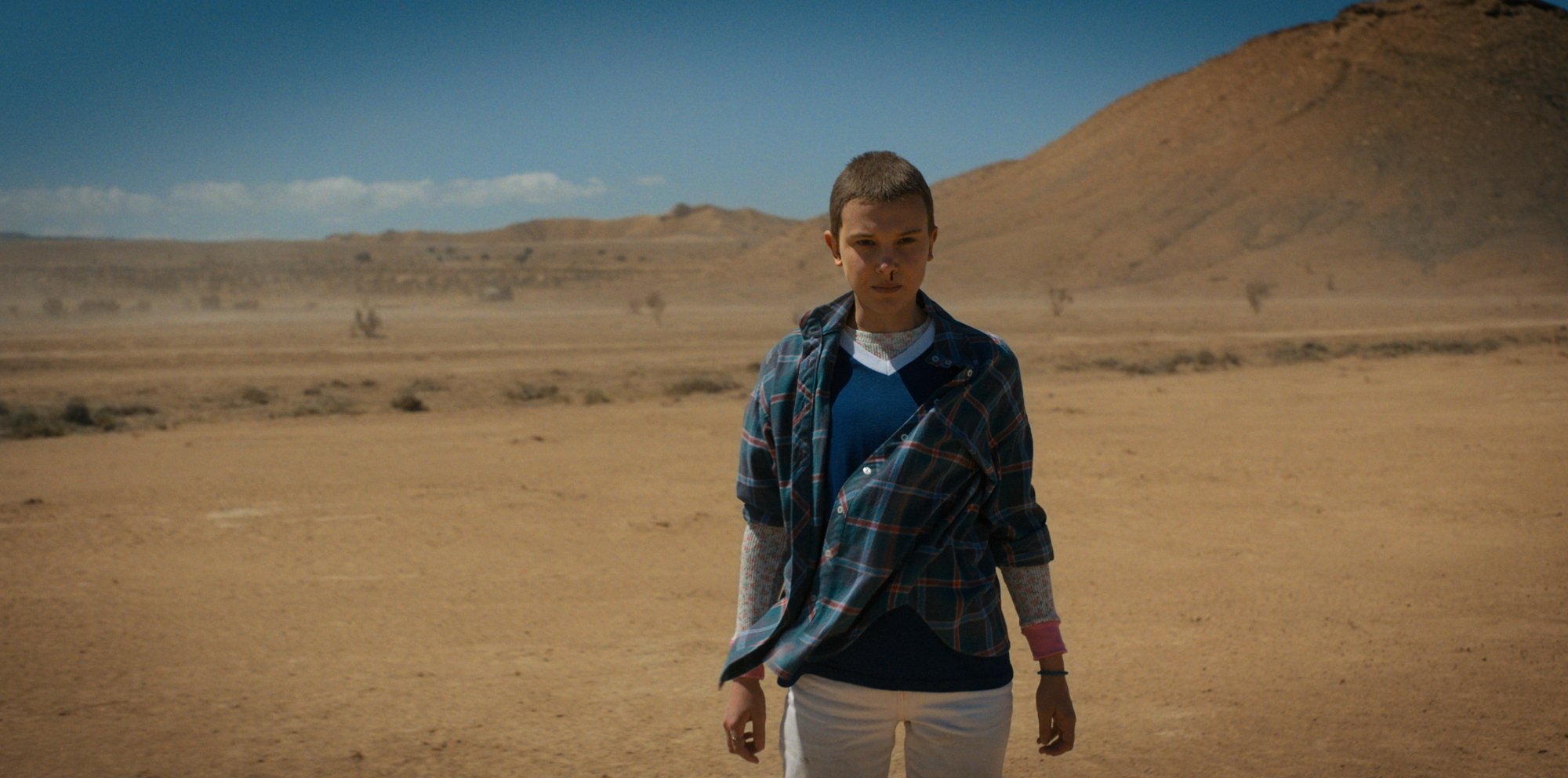 A young woman with a shaved hear stands alone in a desert.