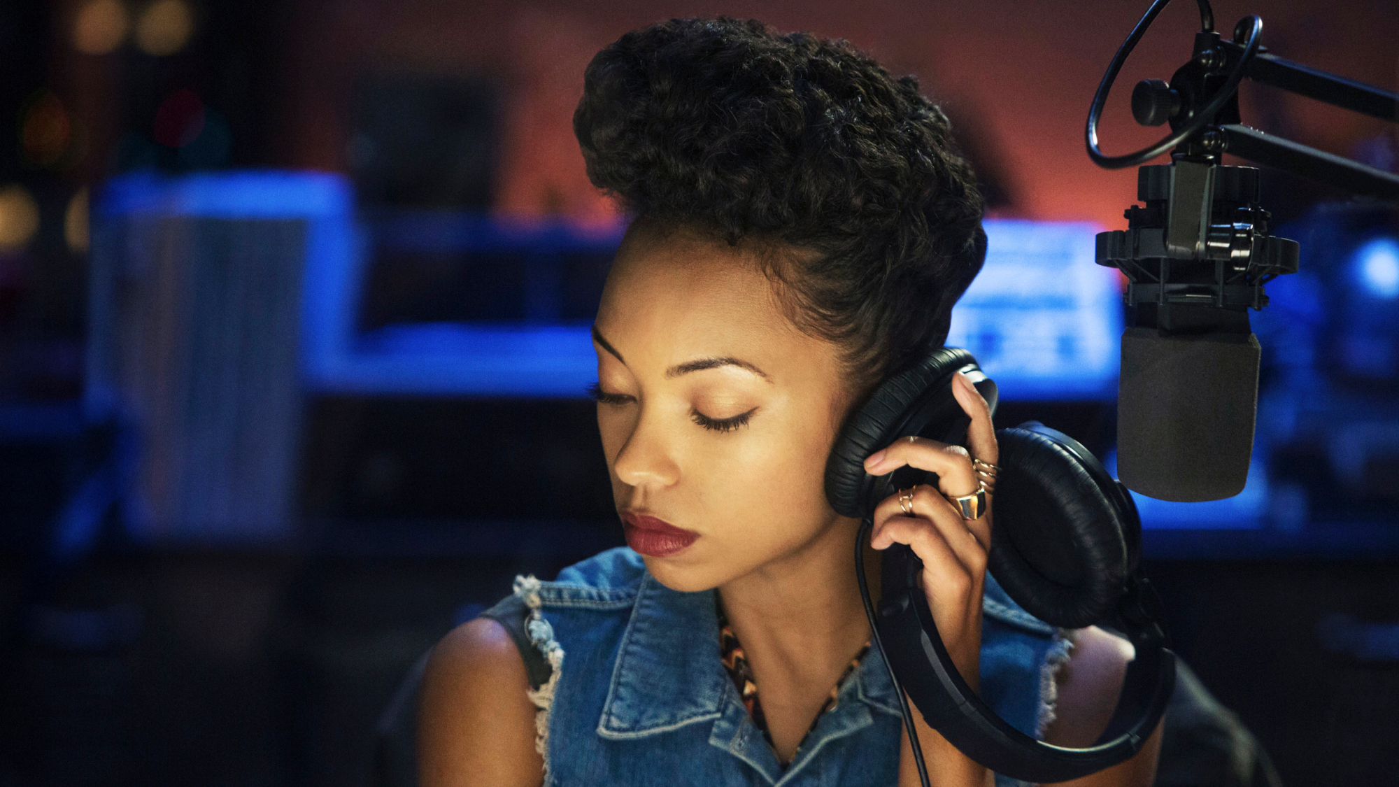 A woman sits in a radio station broadcasting, holding a headphone to her ear.