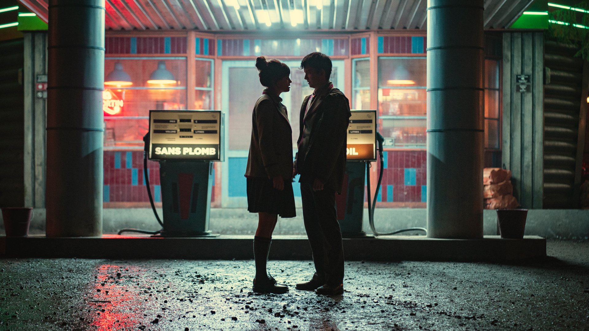 Two teens stand looking at each other at a gas station at night.