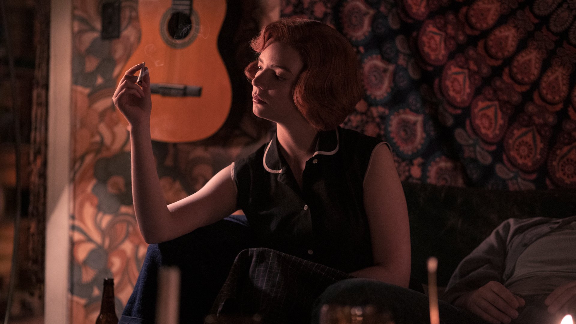 A woman in the 1960s smokes a cigarette in a room where a guitar is mounted on the wall.