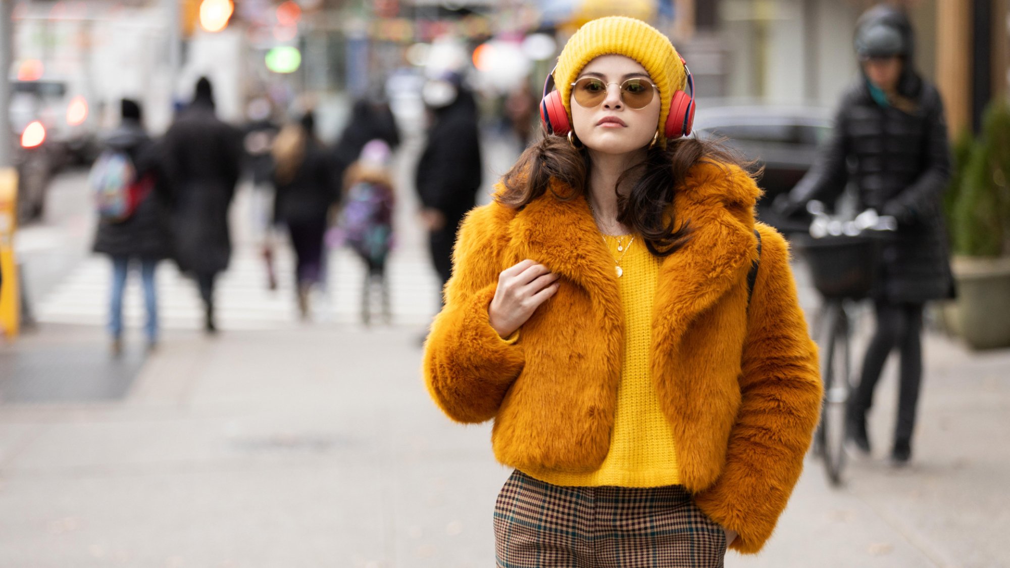 A young woman in a mustard sweater and fluffy jacket with matching headphones and beanie in New York.