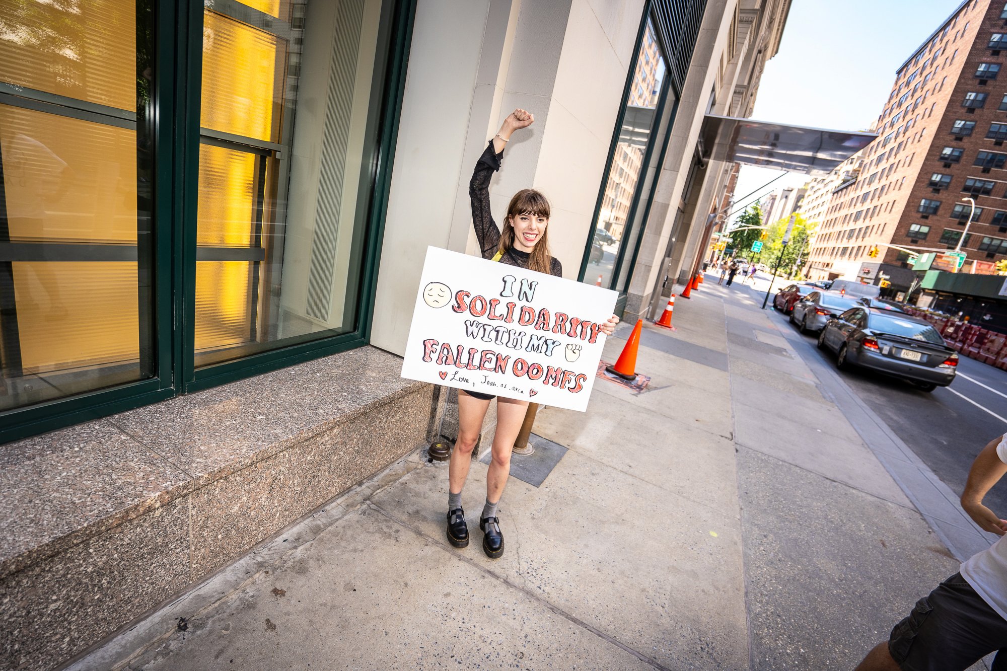 A woman stand with her fist raised holding a sign that says, "In solidarity with my fallen oomfs ."