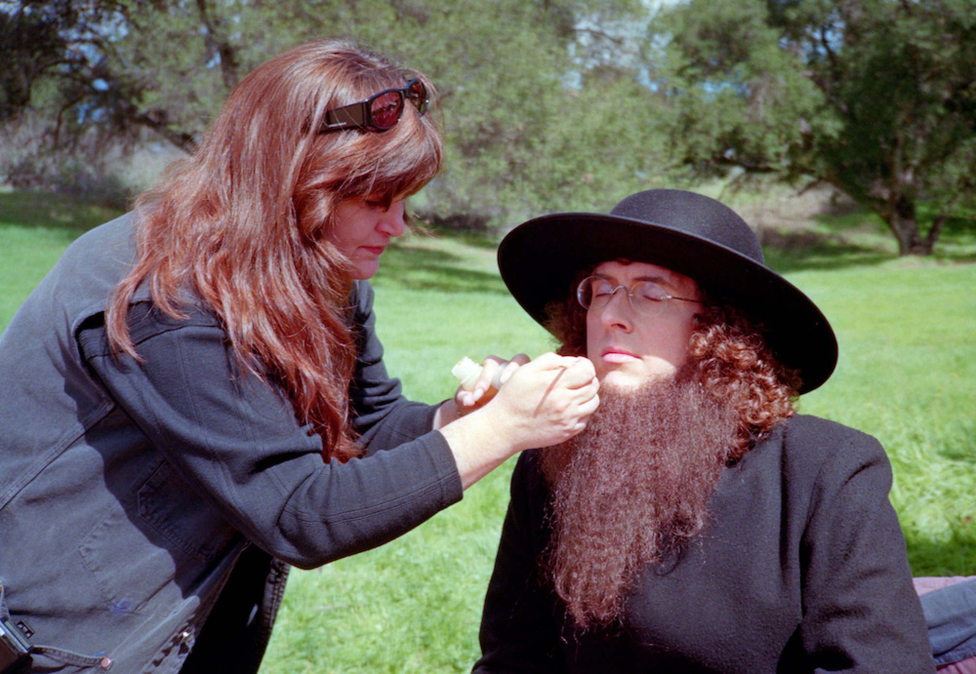 A makeup artist adjusts the beard on a man in all-black attire and a black Amish hat.