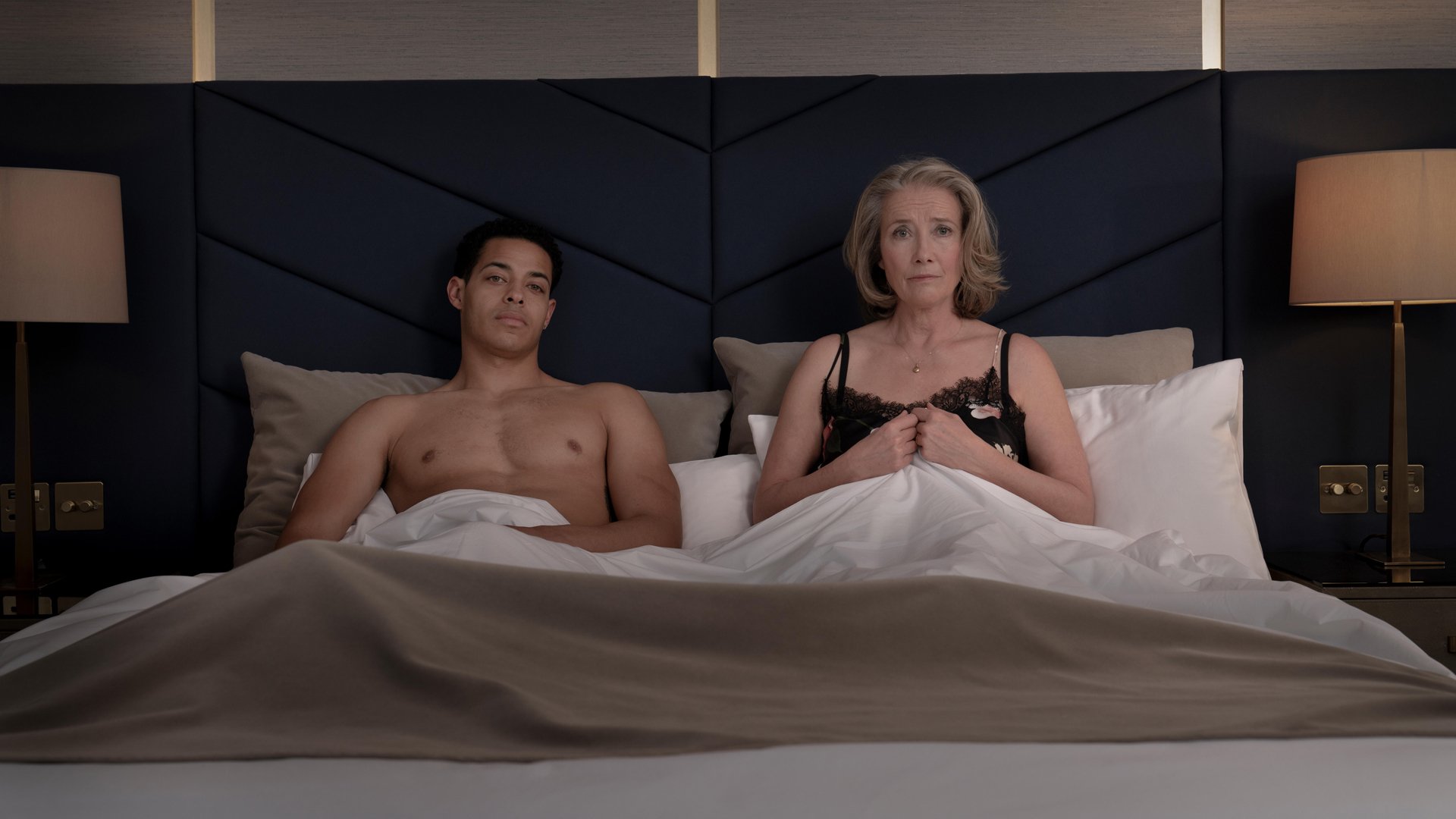 A man and woman in their underwear sit in bed with the covers pulled up, looking at the camera.