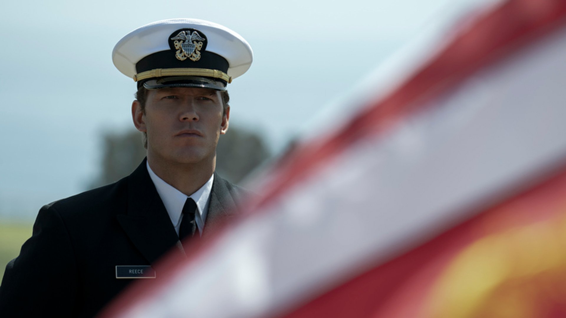 A man in military uniform stands beside an American flag.
