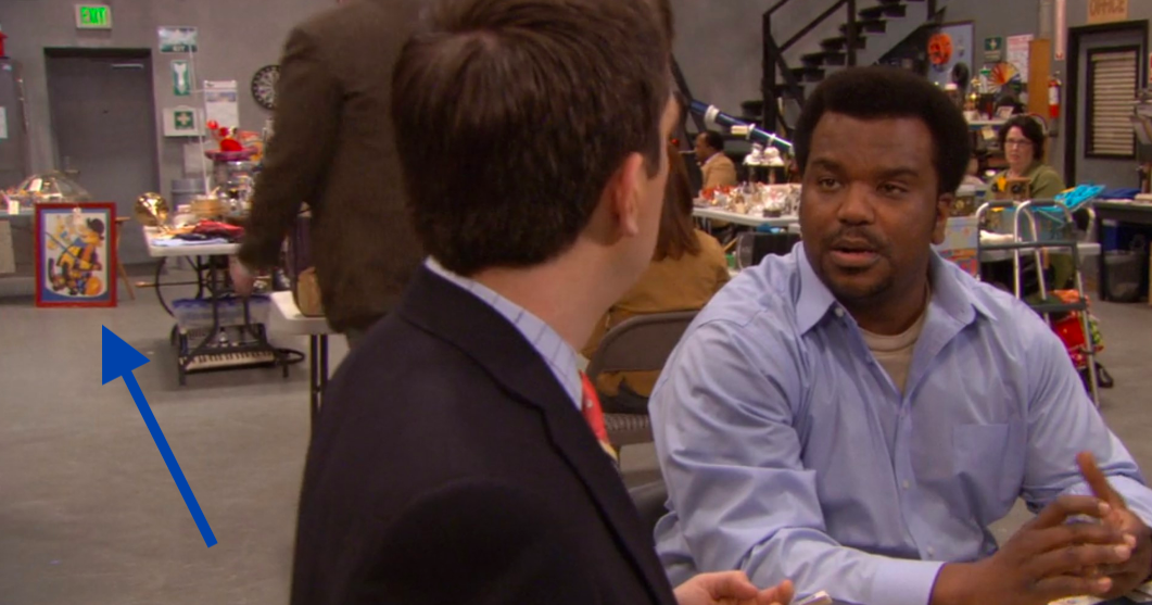 Andy and Darryl chat in the warehouse as the clown painting sits behind them.