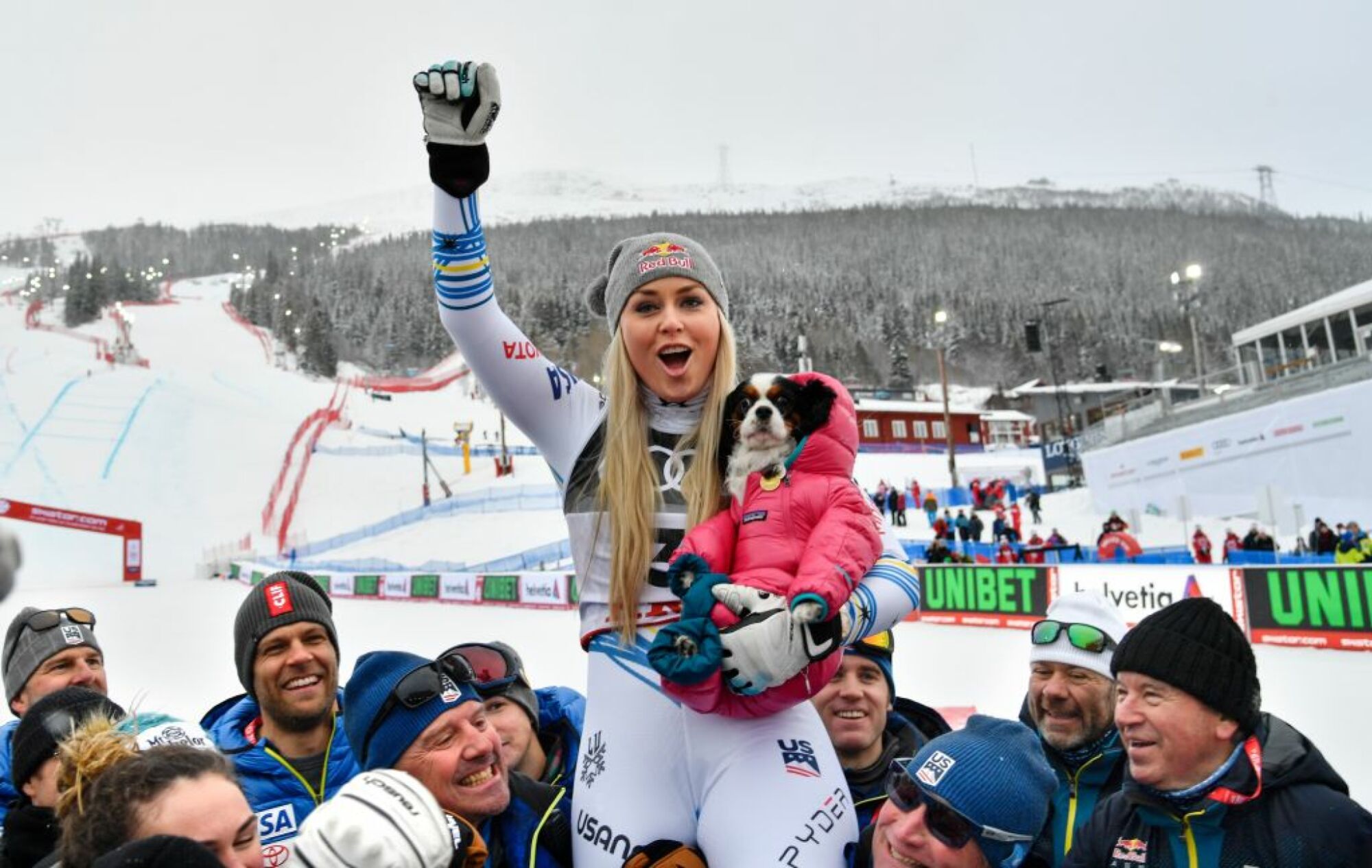Image of Lindsey Vonn celebrating and holding her dog who is wearing a pink puffy jacket