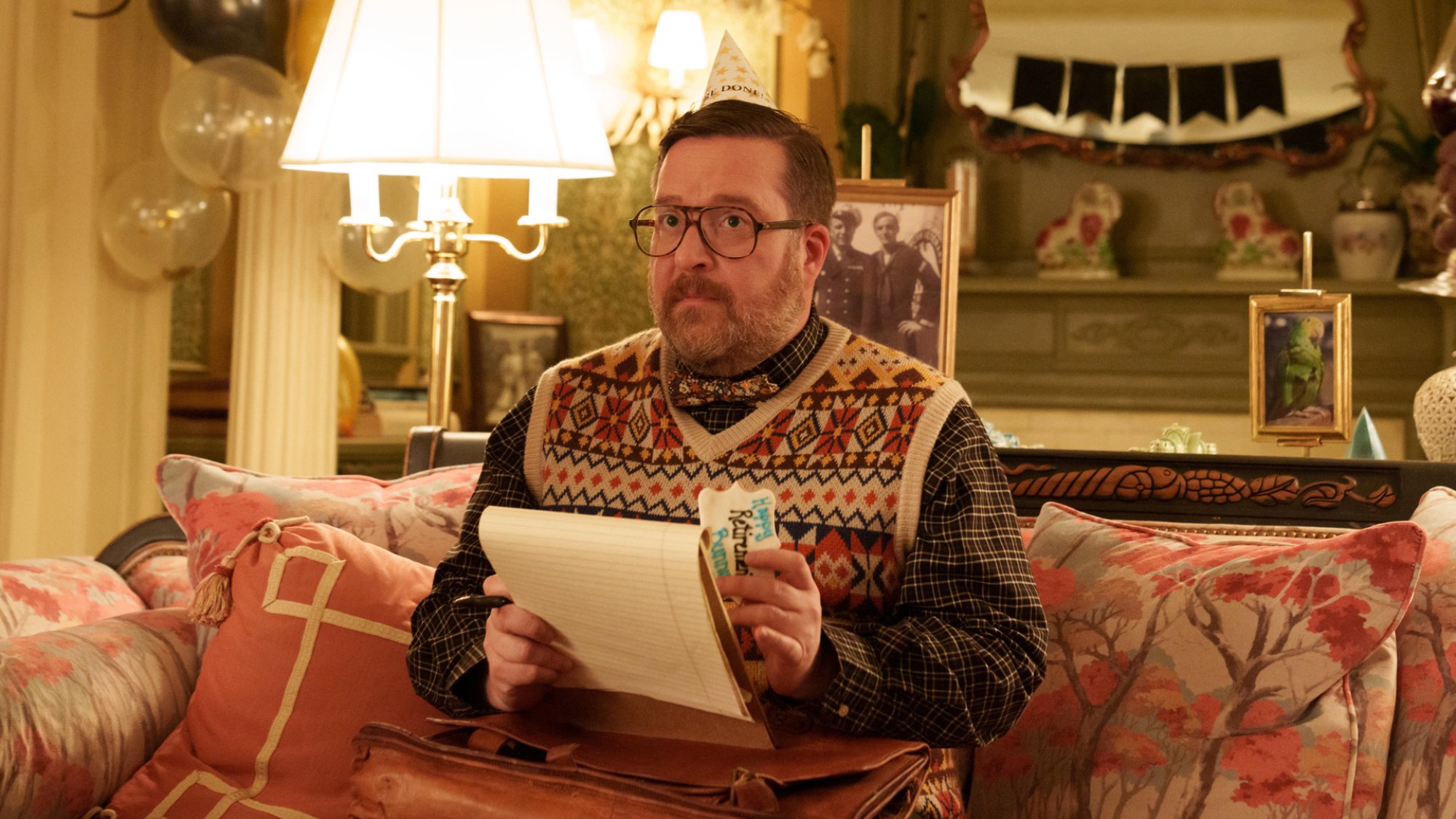 A man in a knitted vest sits in a maximalist apartment living room on a couch.