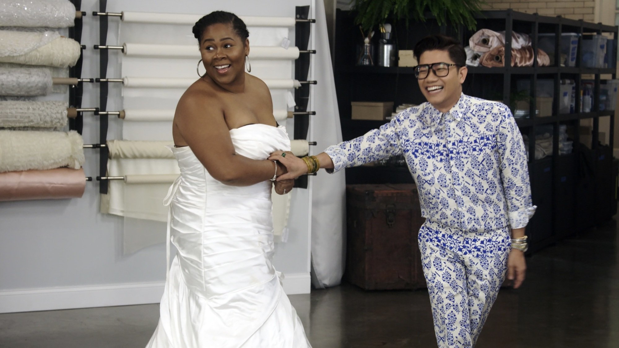 A woman in a wedding dress and a man in a patterned blue and white suit clutch each other's arms in excitement.