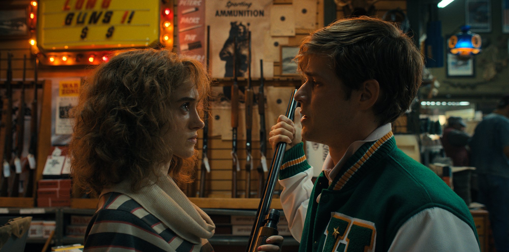 A young woman holds a shotgun in a gun store, and a young man in a letterman jacket holds the barrel.