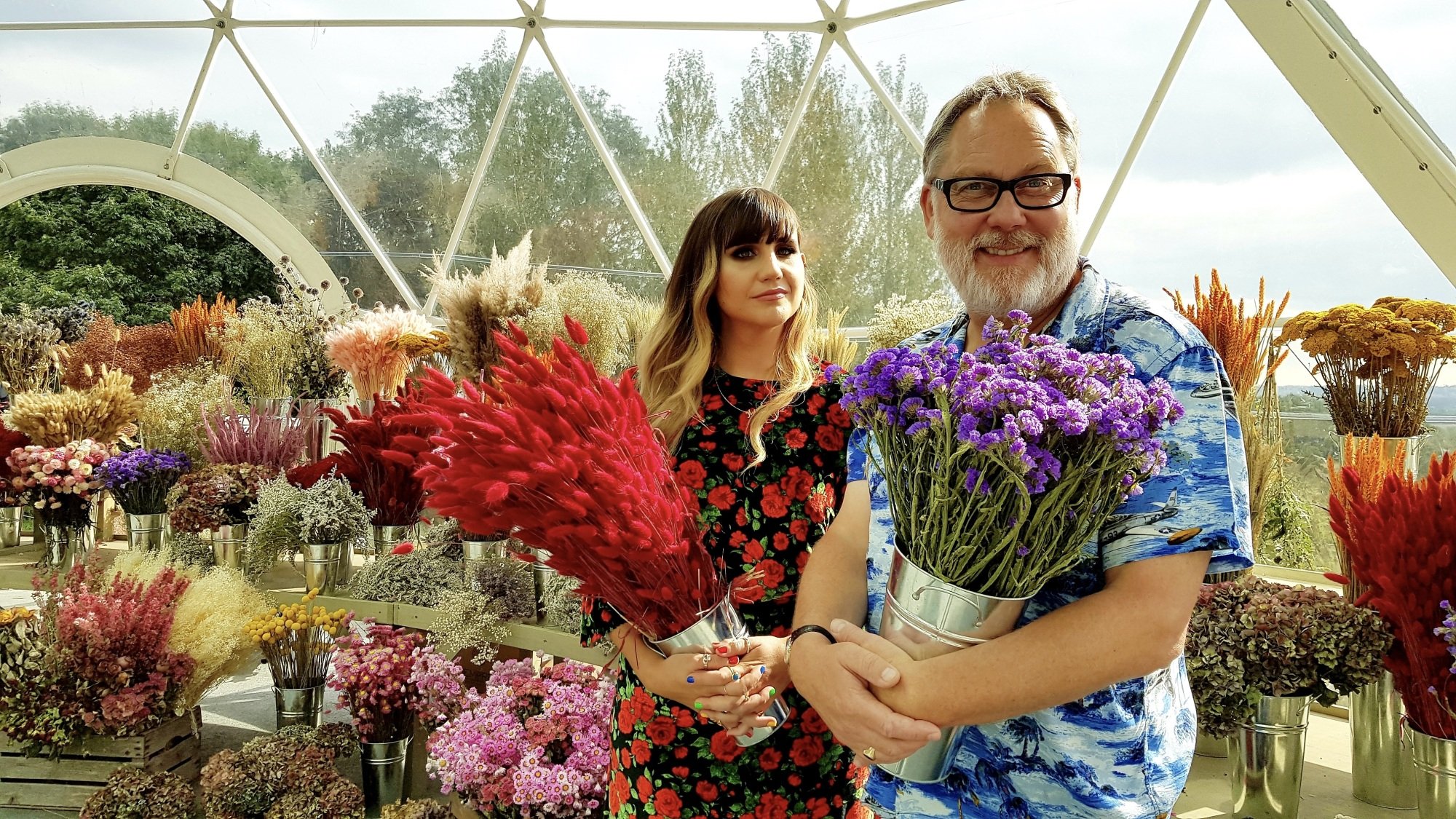 A man and woman hold bouquets of flowers in a greenhouse.
