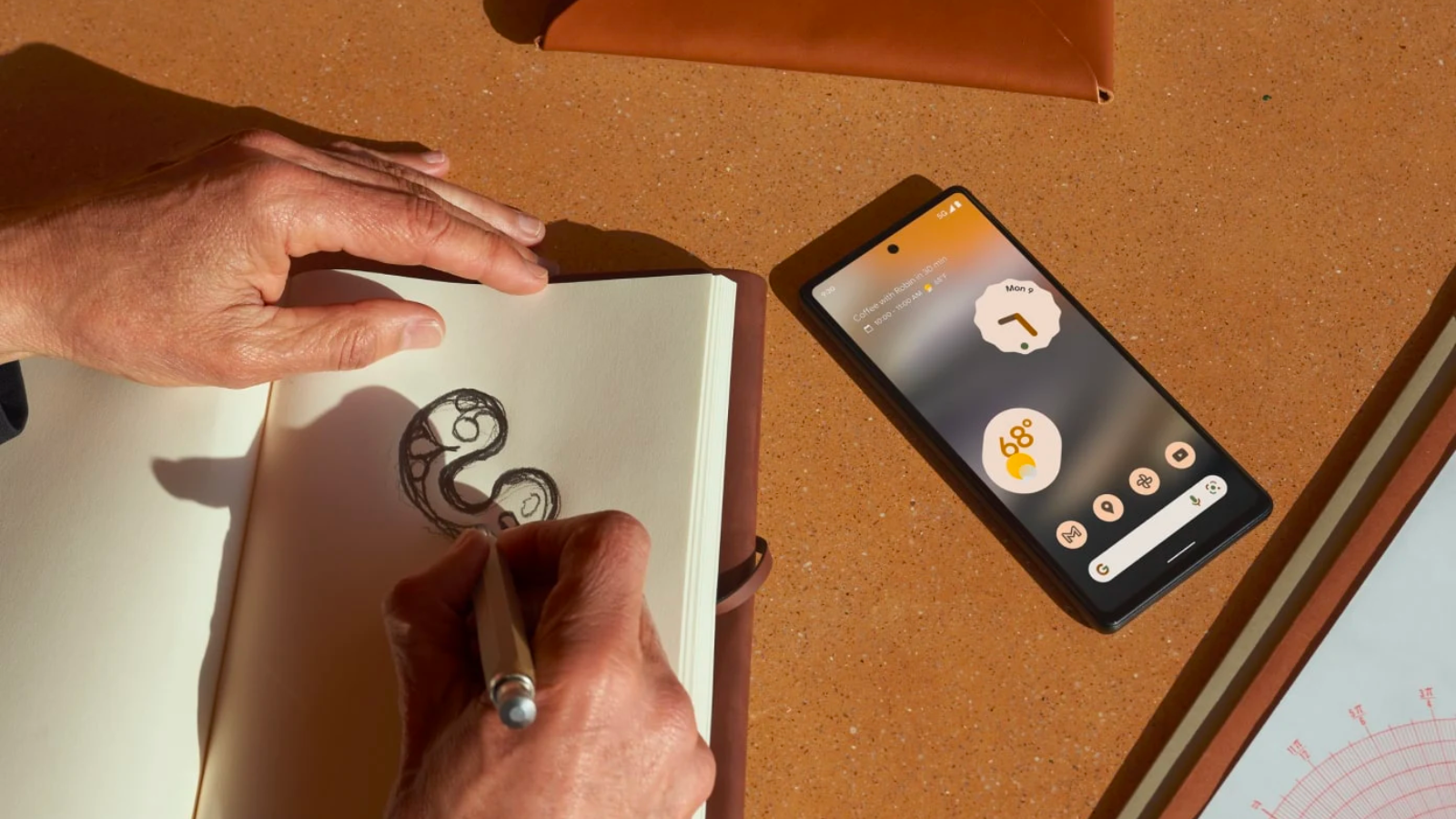a google pixel 6a sitting on a brown countertop next to a person doodling in a leather journal