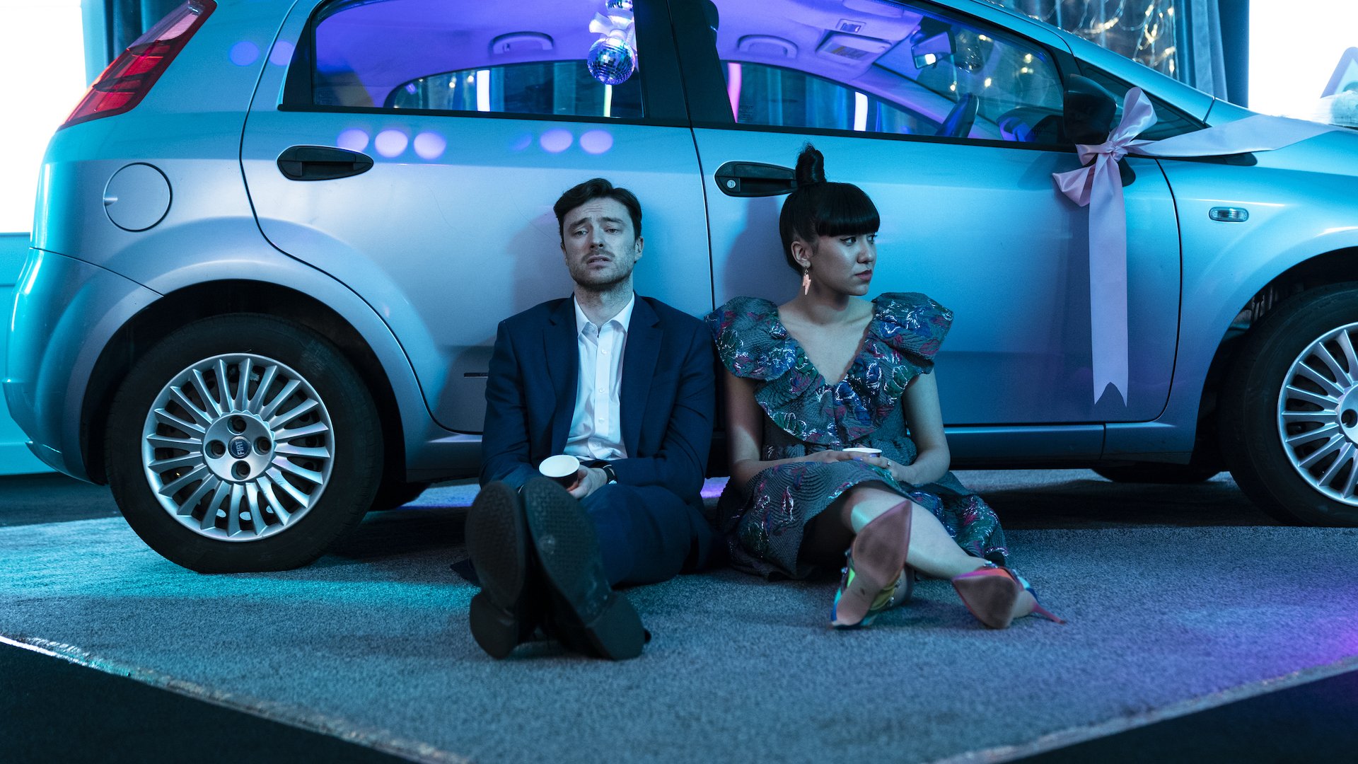 Two people in formalwear sit on the ground beside a car with a bow on it.