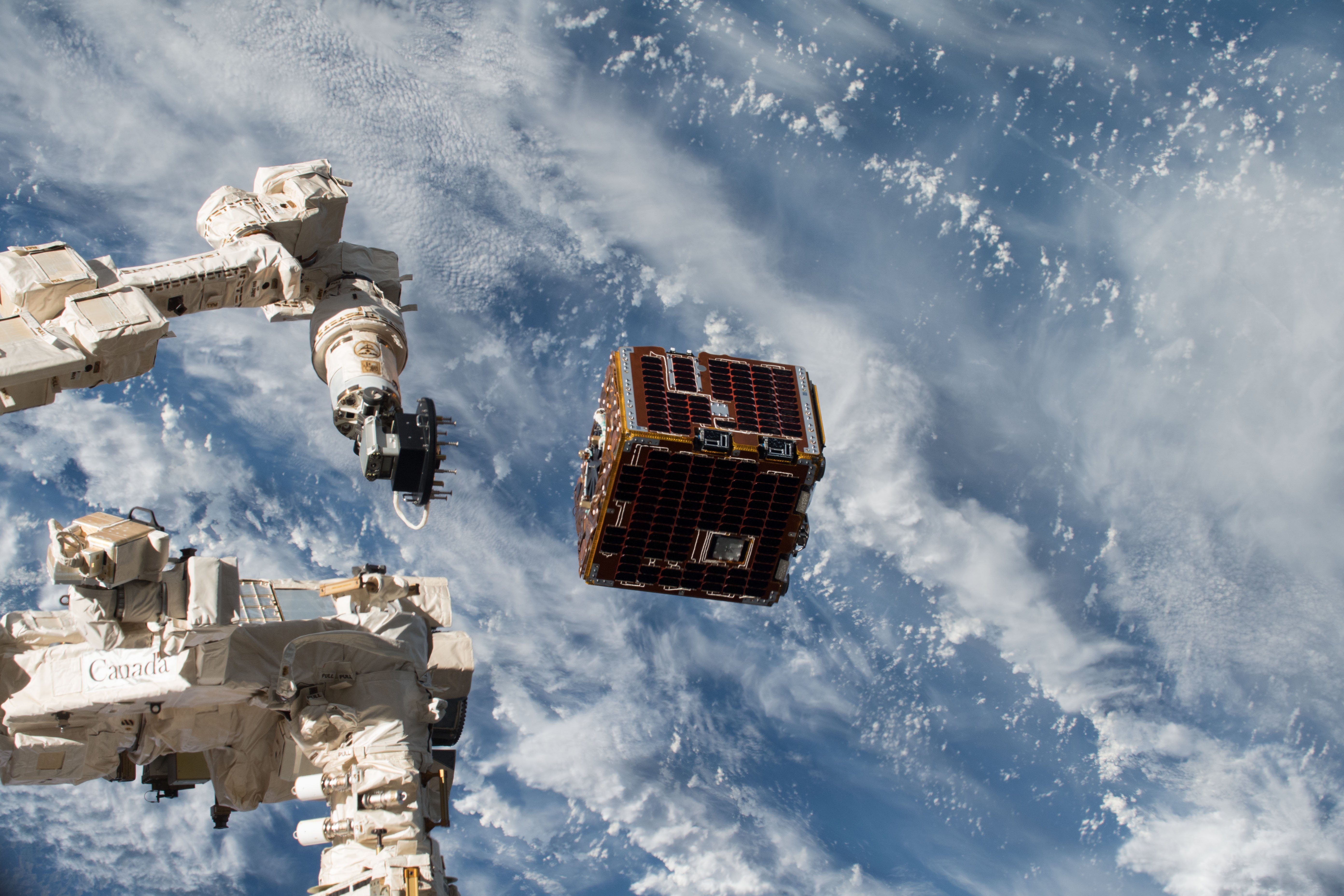 A red and black metal cube floats in space next to a white satellite arm. Behind the two objects is the Earth's cloud-filled atmosphere.