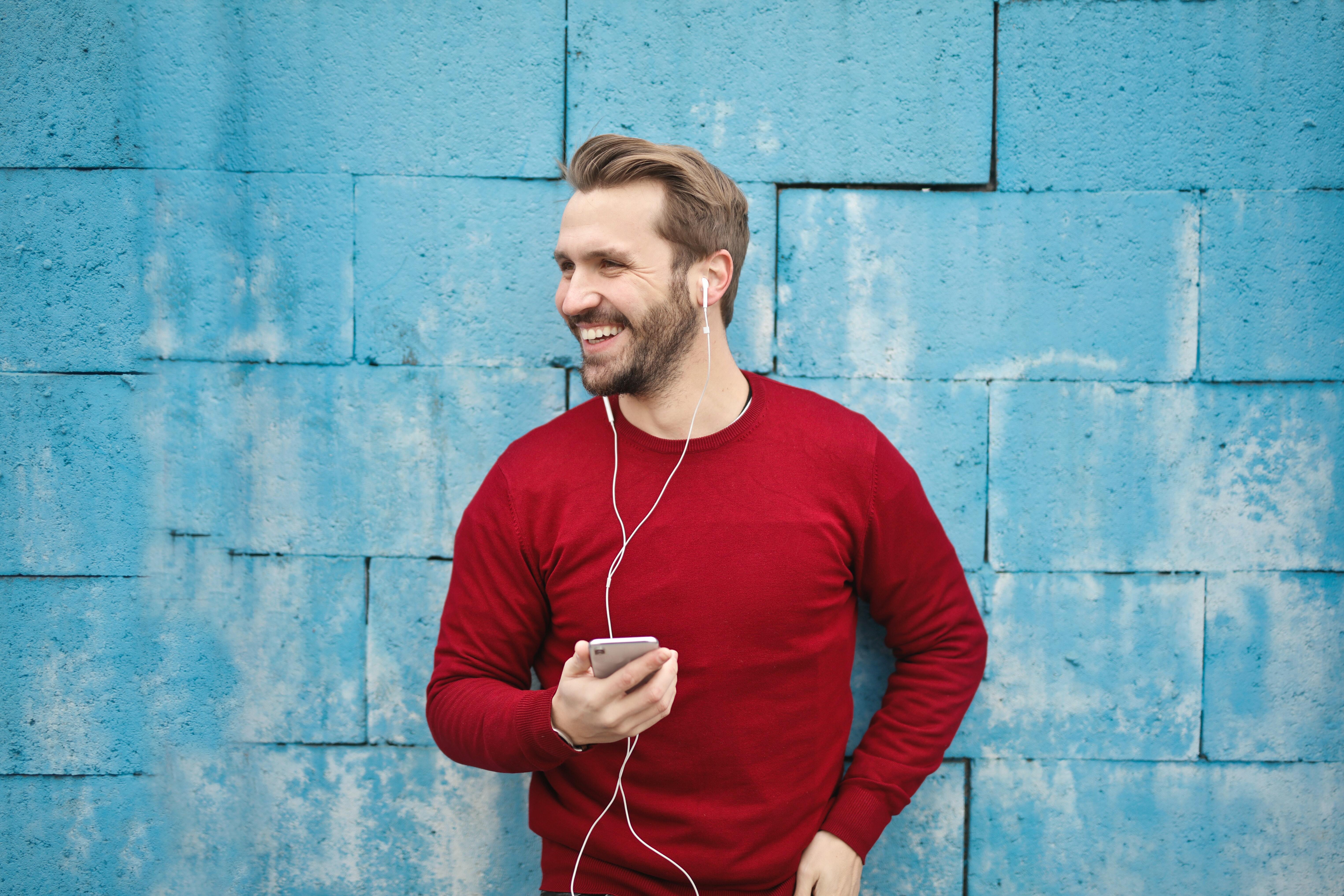 A man in a red sweater listening to earphones, standing in front of a blue wall.