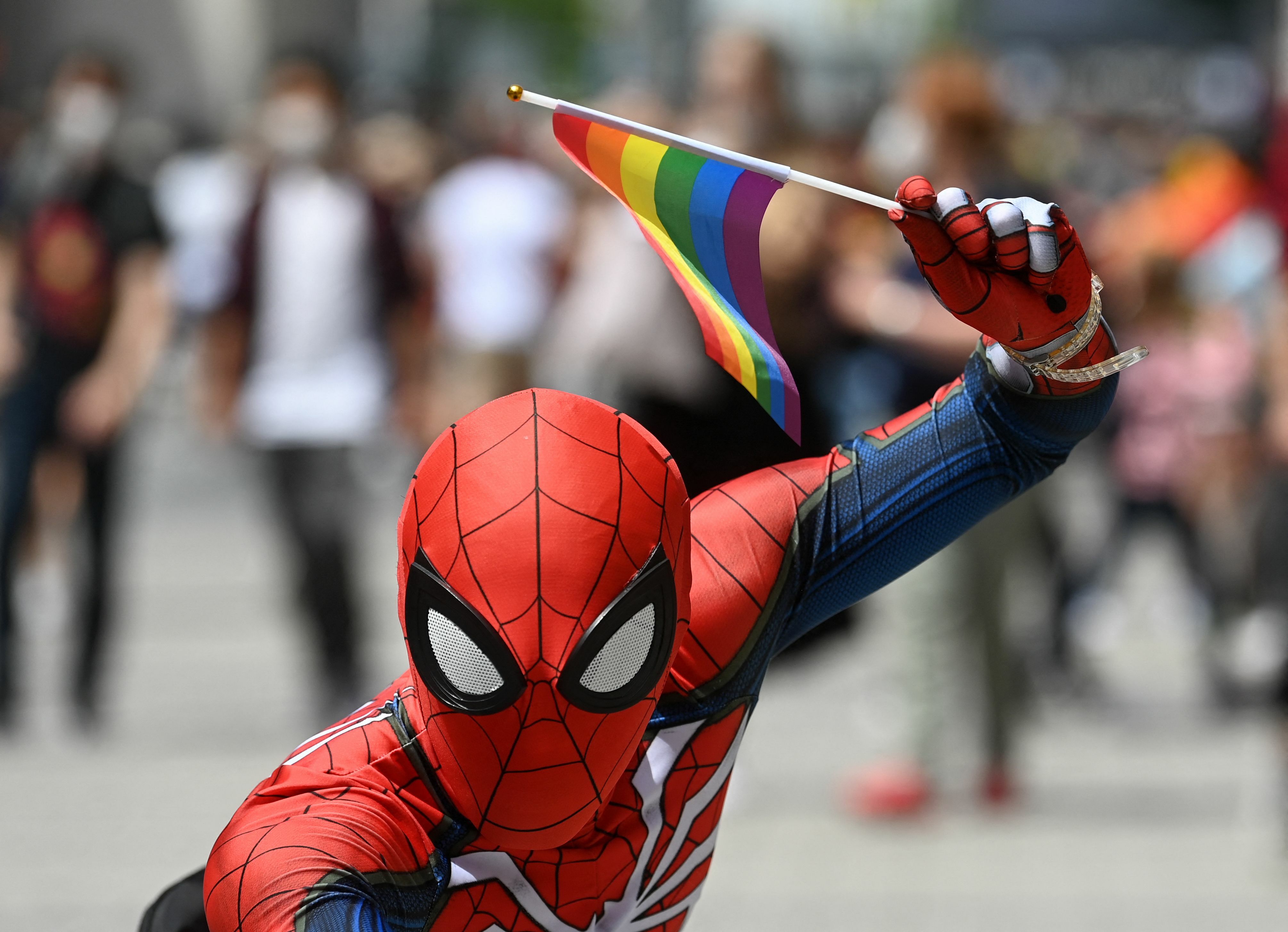 An young man dressed up as comic book character Spider-Man holds up a rainbow flag