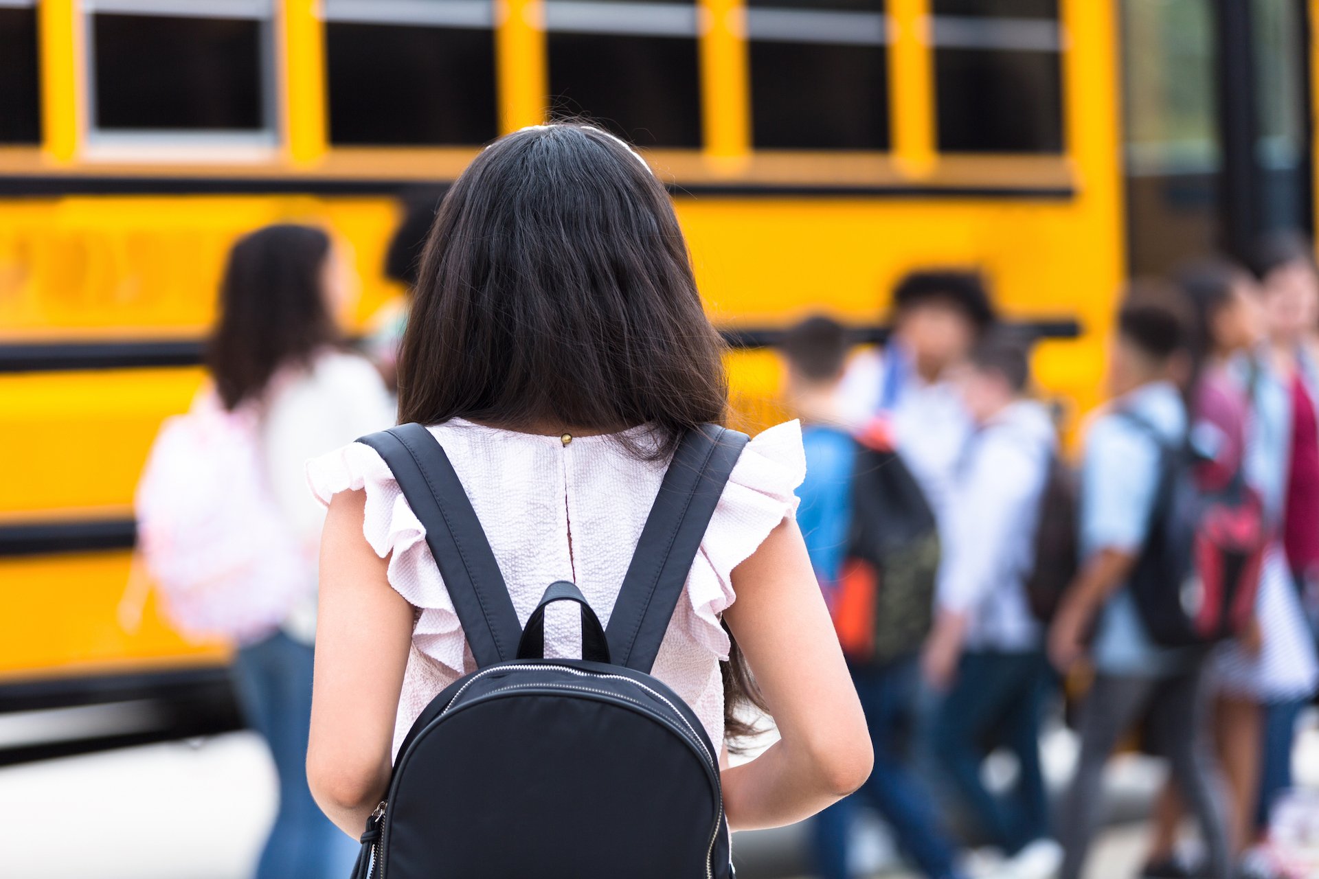 A girl wearing a backpack tentatively approaches a school bus with students waiting to board. 