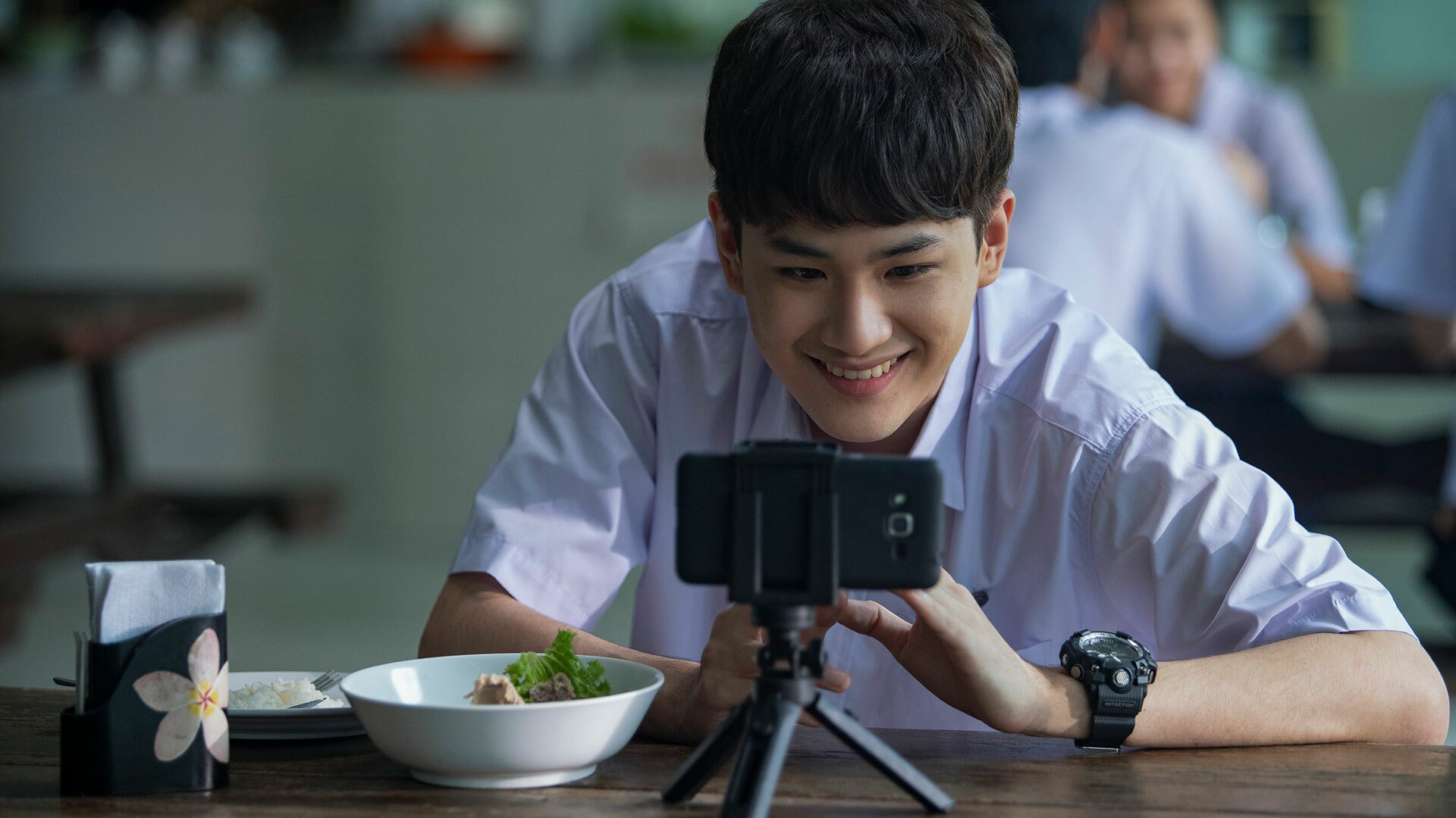 A boy sitting in a lunch cafeteria smiles at his smartphone, which rests on a tripod.