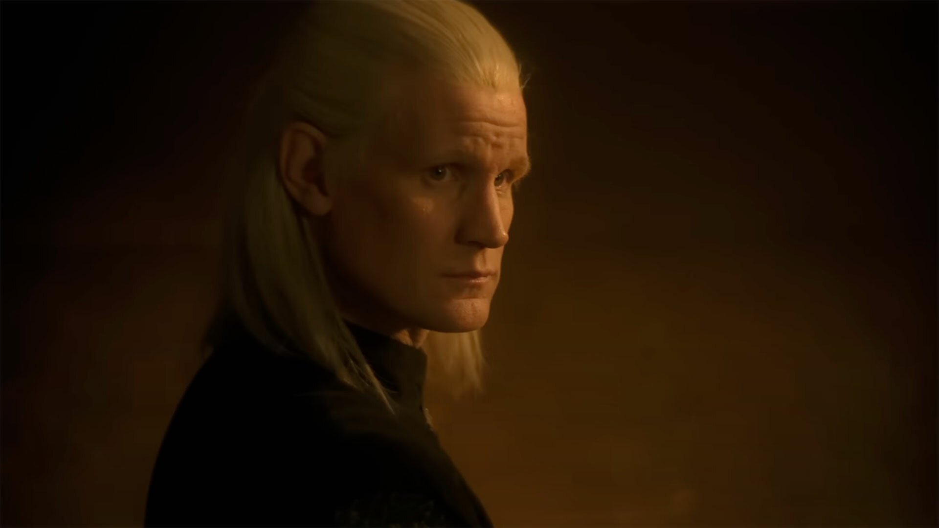 A man with long blonde hair stands in a dark room.