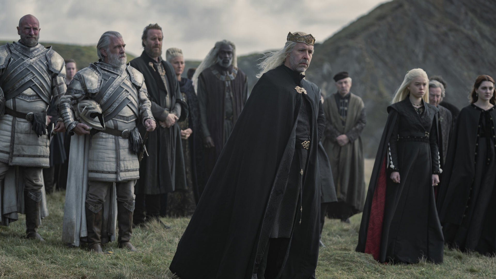 A group of people in mourning clothes, including a King and some knights, stand on the coast.