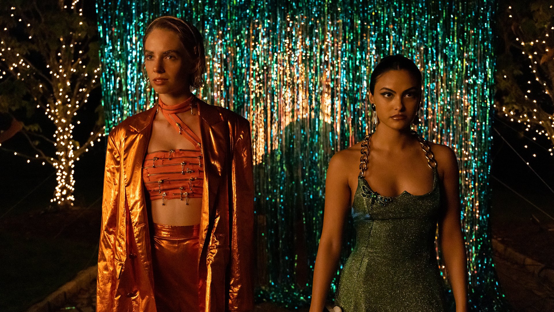 Two young women in party wear stand in front of a shimmering wall.
