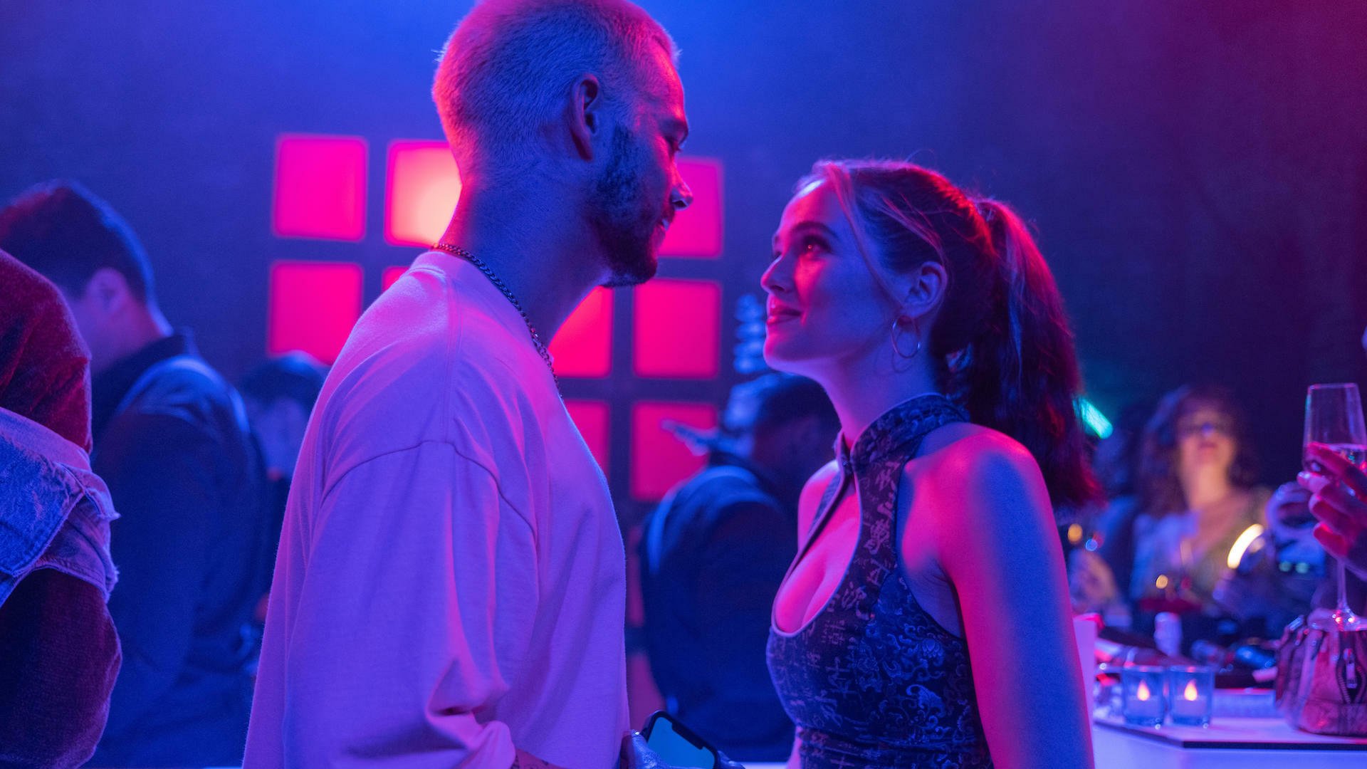 Two people gaze at each other at a party bathed in pink and purple light.