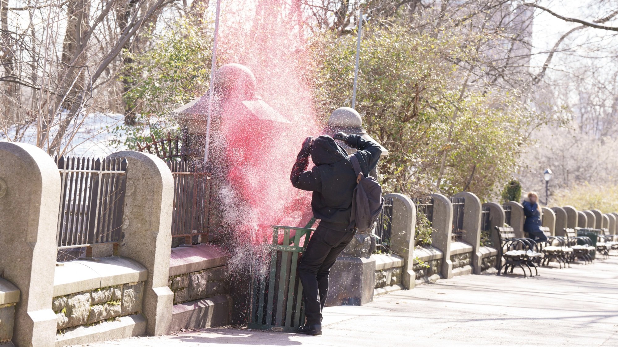 A man all in black recoils from a park trash can with red glitter exploding from it.