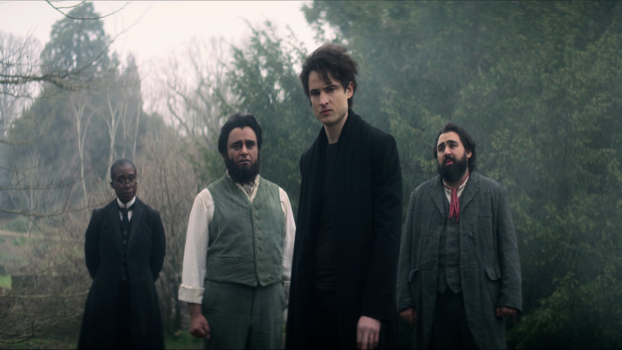 Four people in formal attire stand in a misty cottage yard looking sombre.