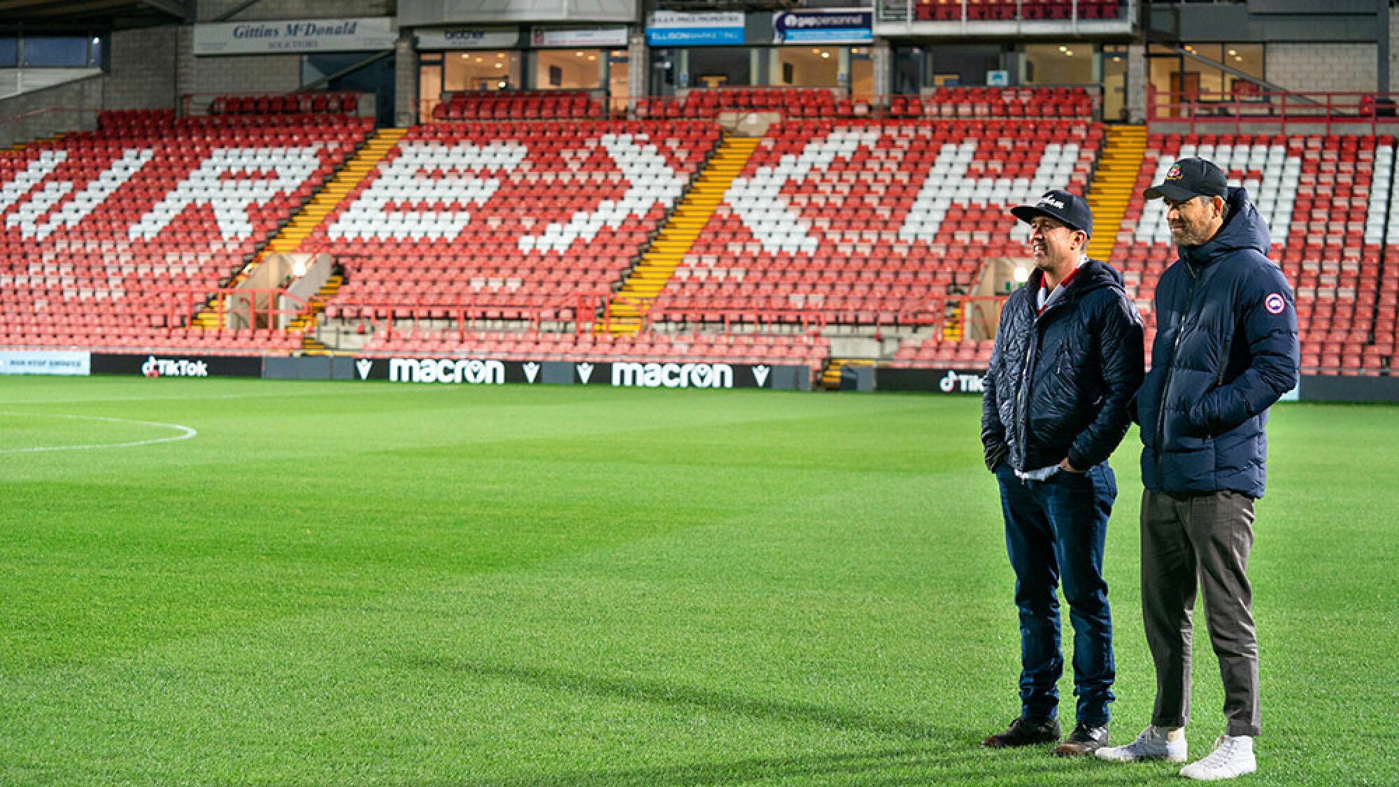 Two men stand on the empty pitch of a football stadium, with the word "Wrexham" spelled out on the seats in the background.