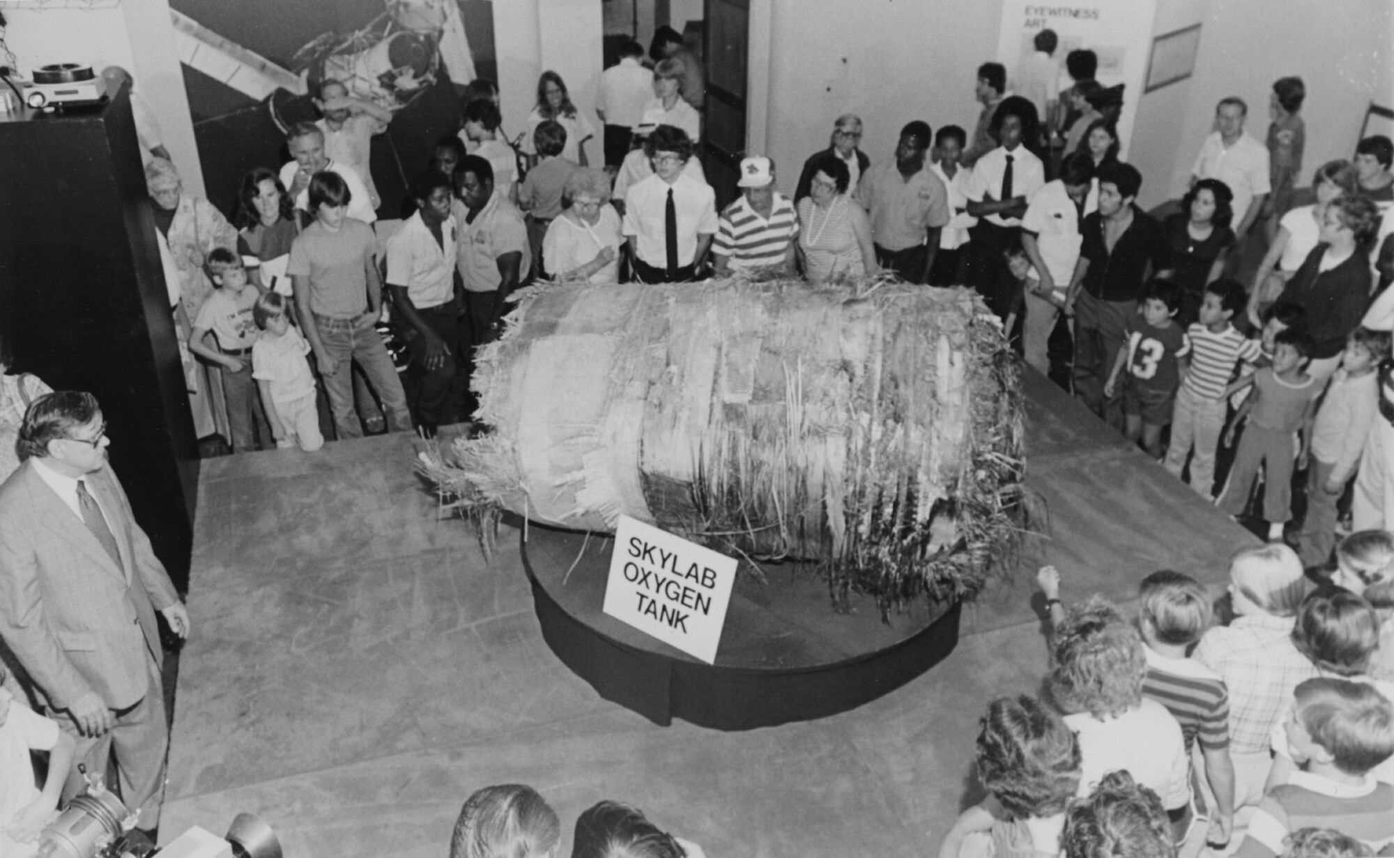 A black and white photo from 1979. A group of museum visitors gather in a circle around a large cylinder-shaped piece of shredded metal. It sits on a round pedestal with a sign that reads "Skylab oxygen tank". 