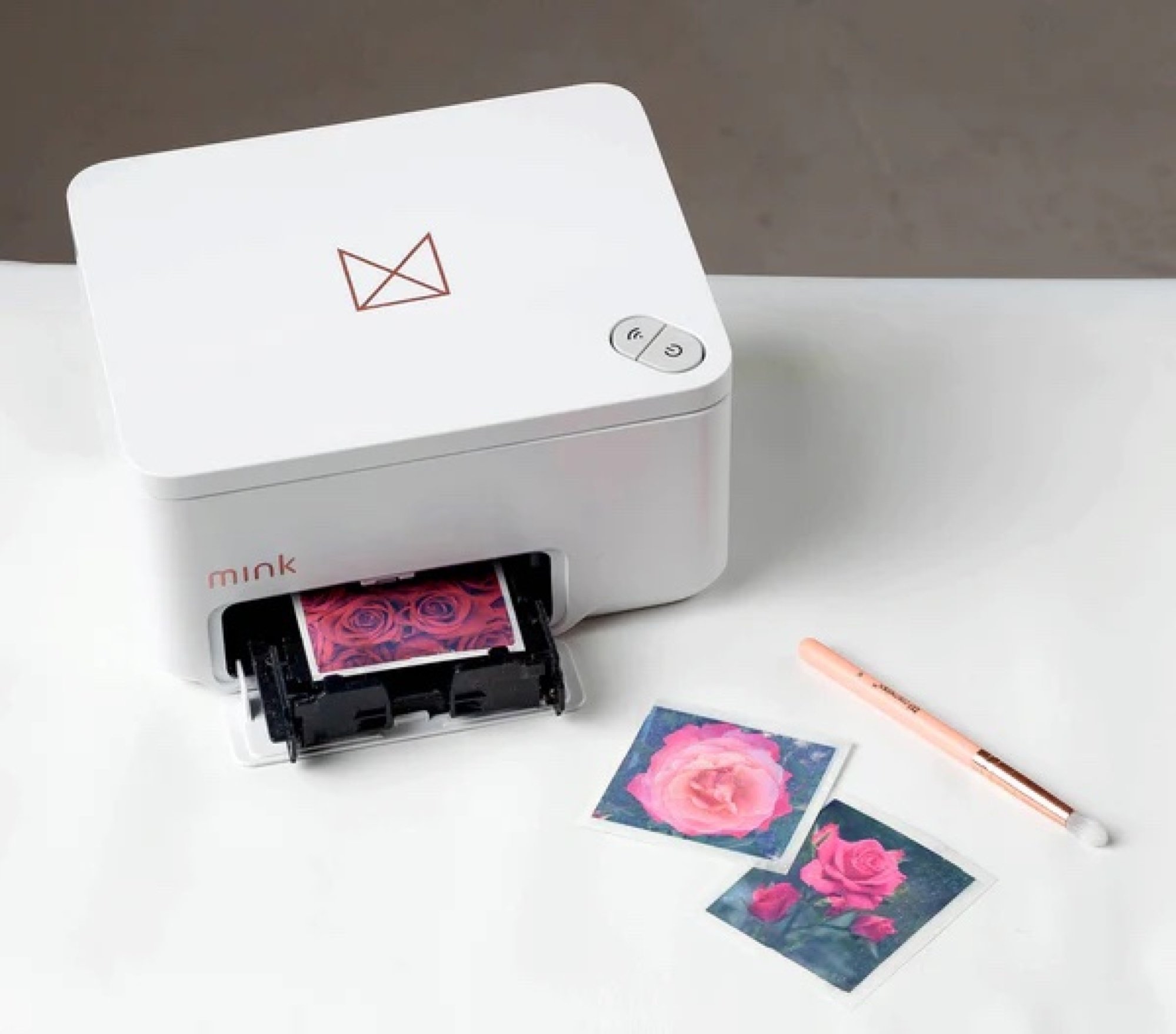 Small white printer on table top in the middle of printing out a square image of a pink flower. There is a makeup brush next to the printer.