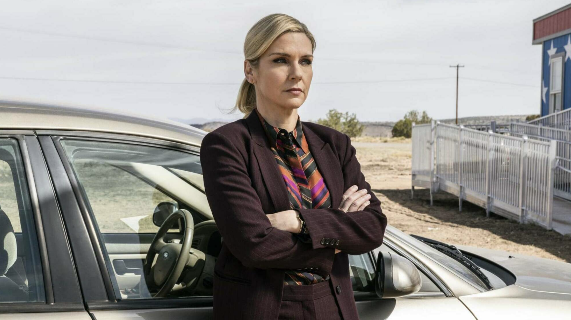A woman in a suit leans on a car in the desert.