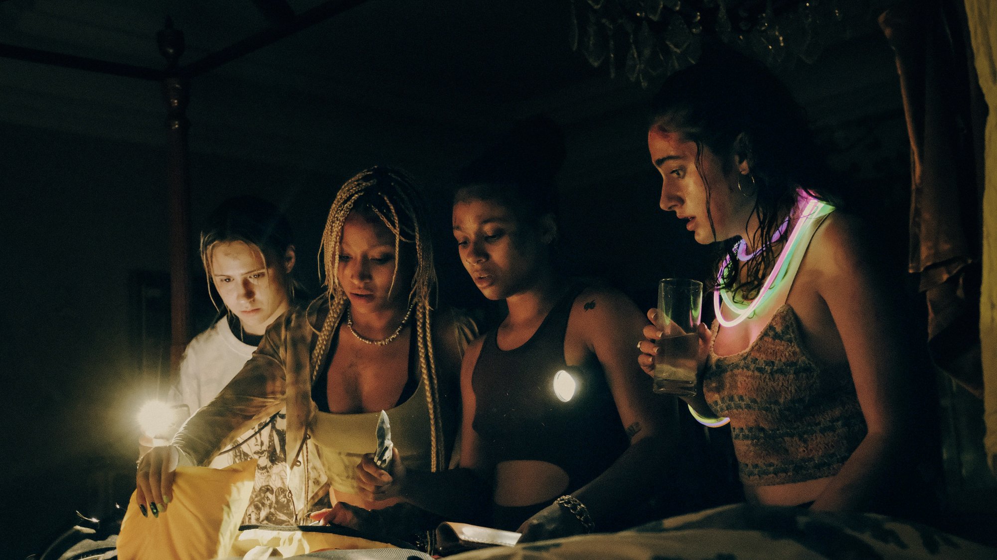 Four young women gather around a bed, shining flashlights on an open suitcase.