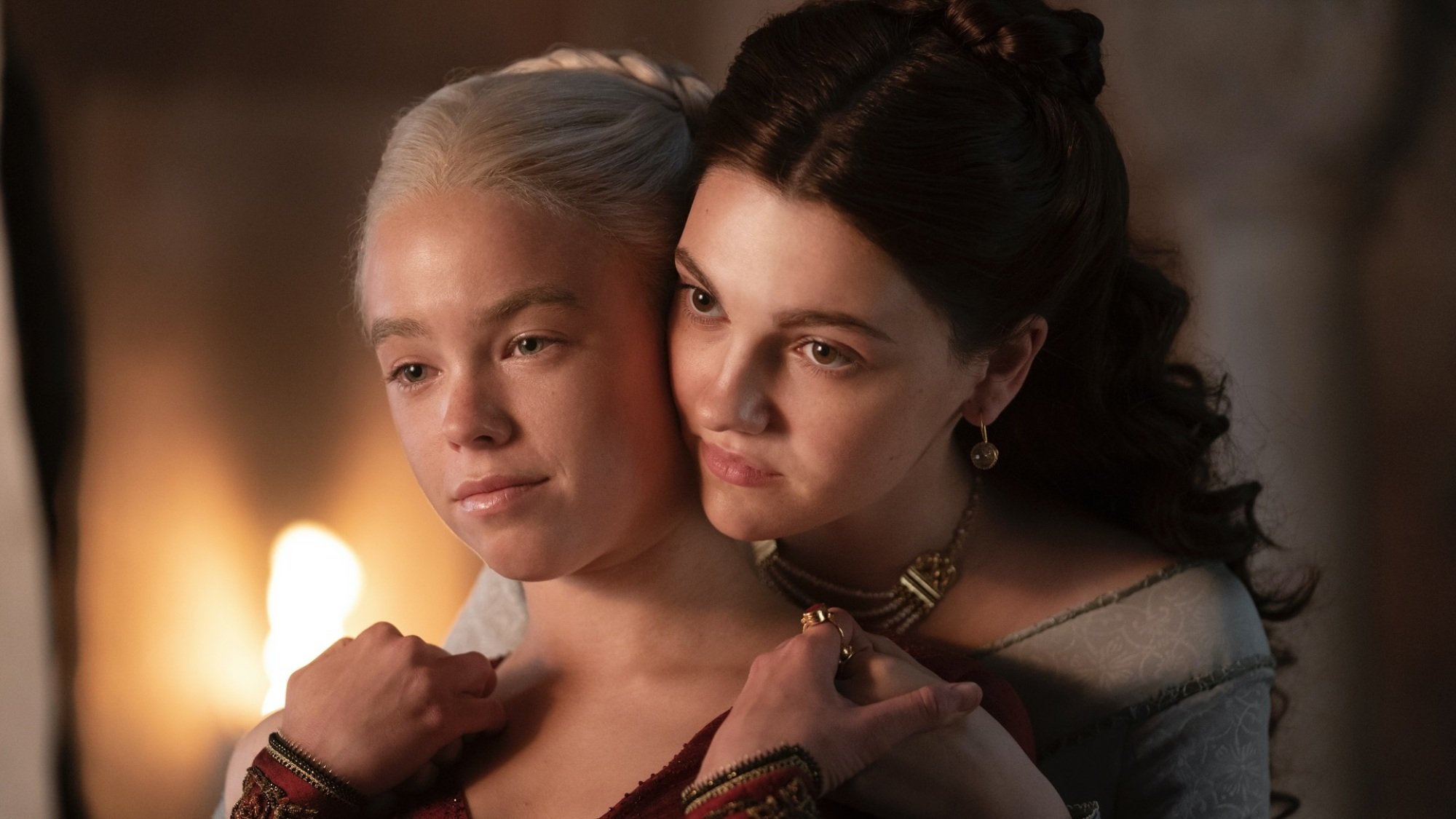 Two young women in lavish fantasy dresses. One stands with her back to the other, but they're holding hands over each other's shoulders.