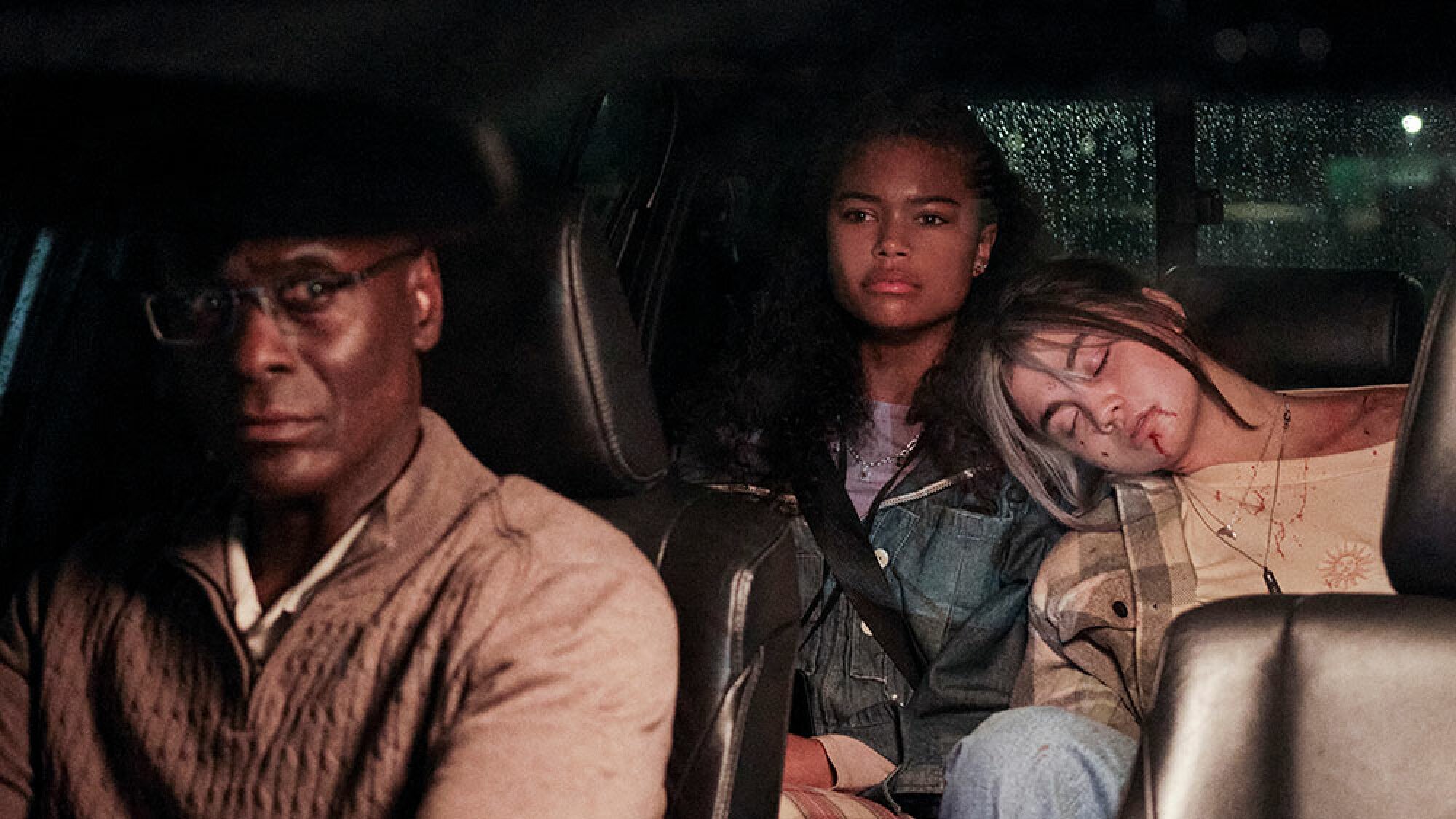 A man with glasses drives two teenage girls, one of whom rests her head on the other's shoulder in the back seat.