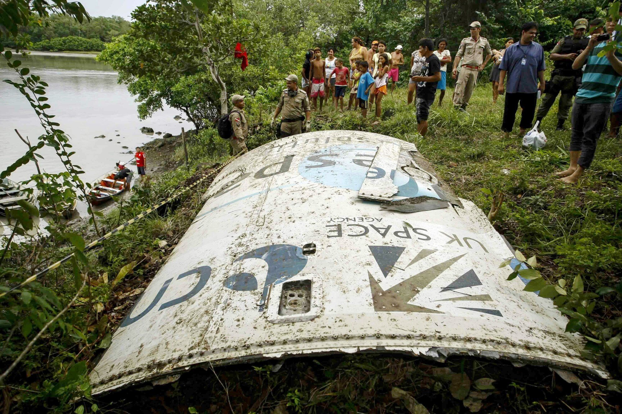 A large, white piece of debris from a spacecraft rests in the grass on the bank of a river in Brazil. The piece of metal has the words "UK space agency" printed on it. A group of people stand behind the debris.