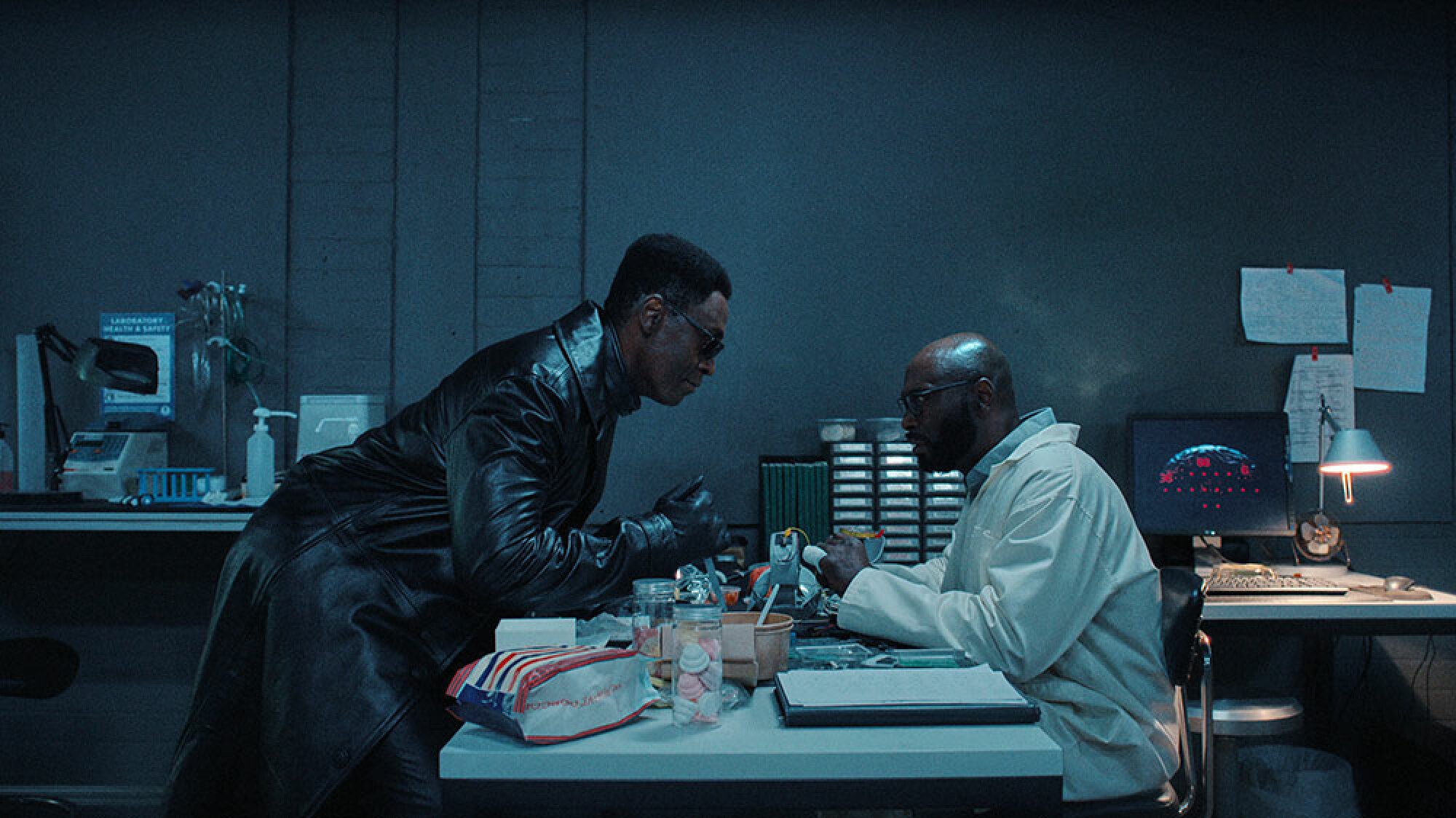 A man in sunglasses and a leather coat hovers over an identical man wearing glasses and a lab coat.