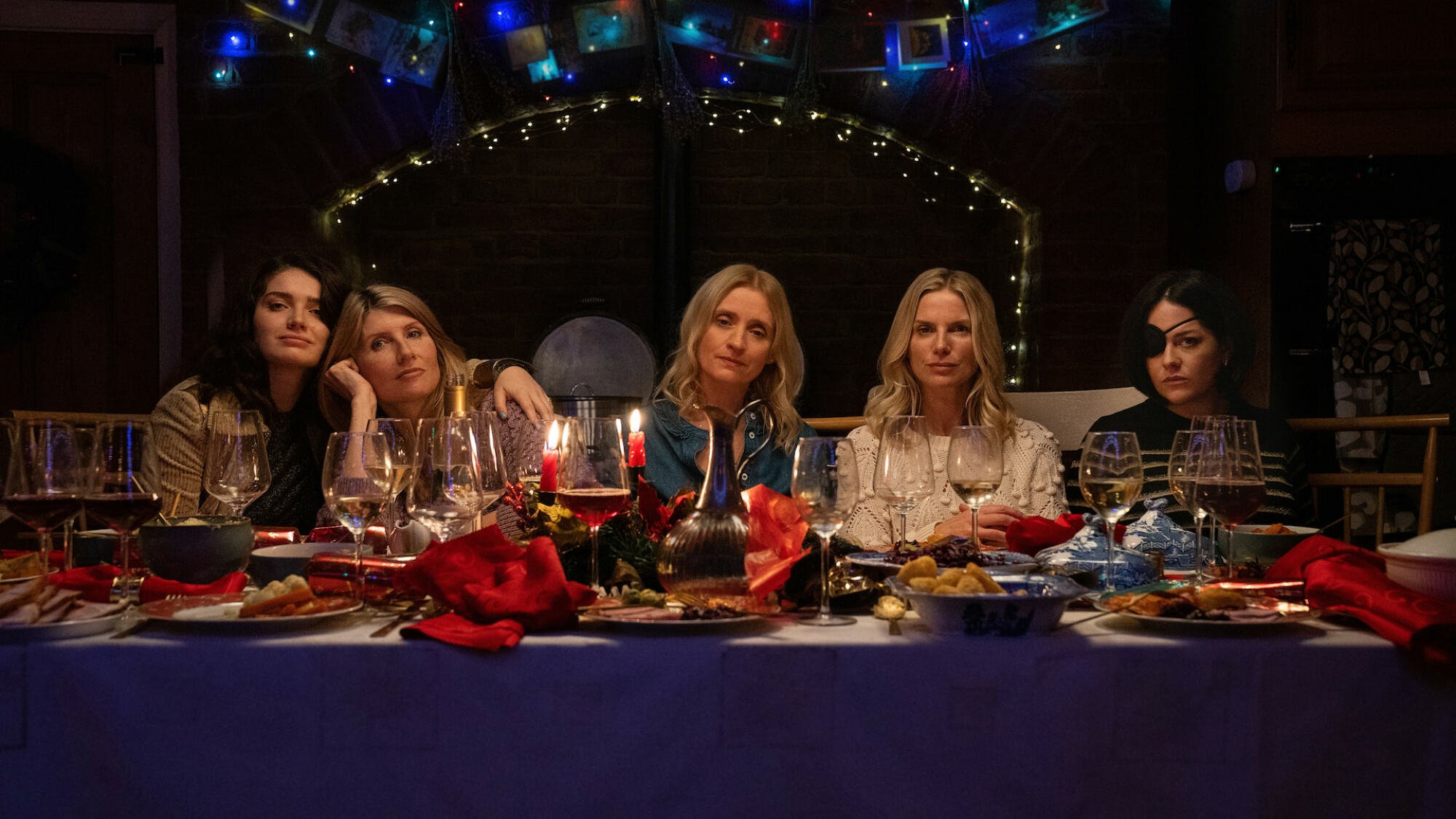 Five woman sit at a festive dining table looking at the camera blankly.
