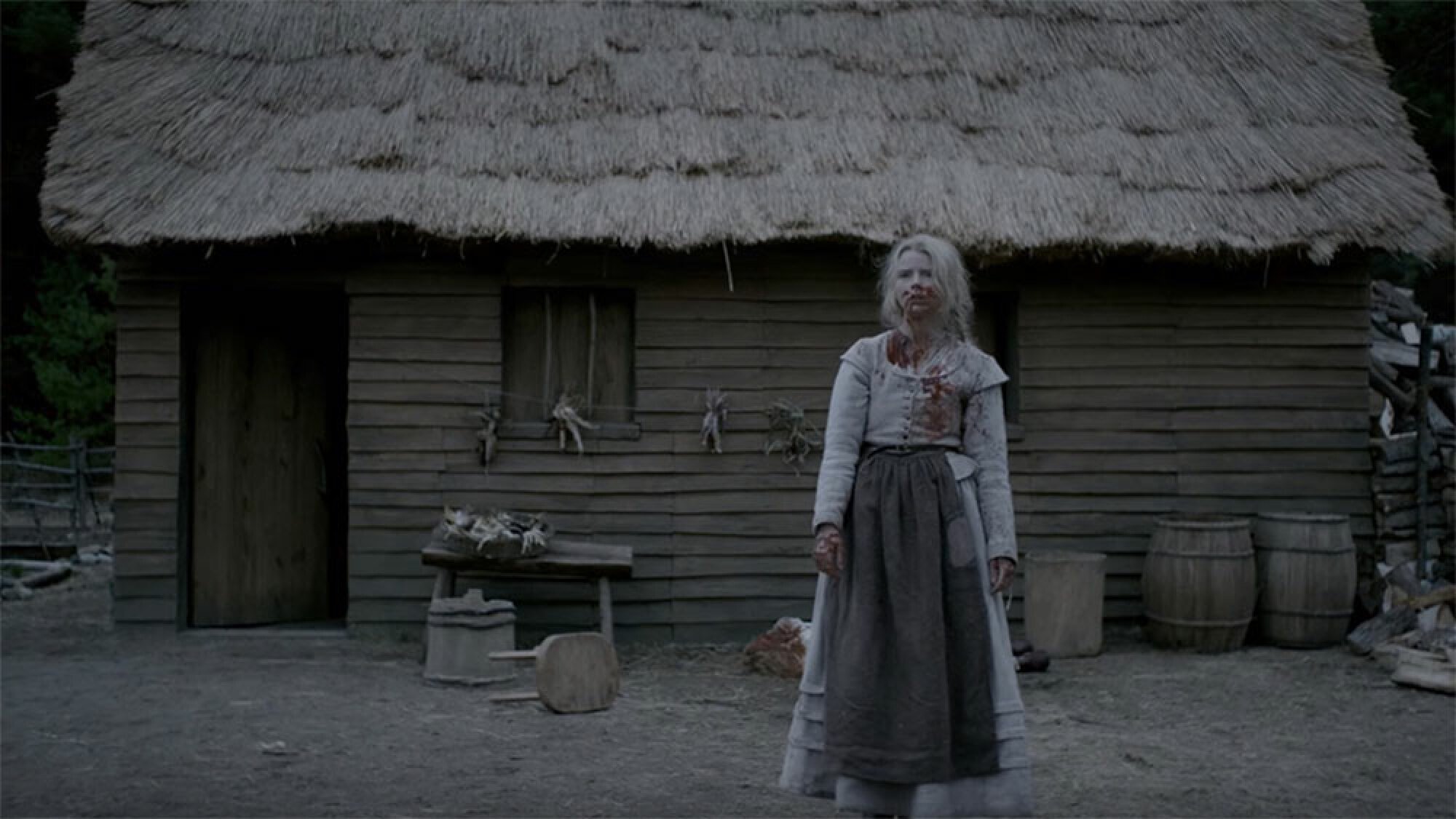 A woman covered in blood stands in front of an old thatched building.