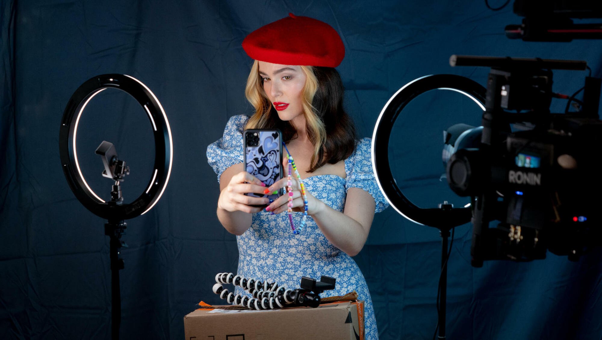 A woman in a red beret and blue dress takes a selfie on her phone with ring lights.