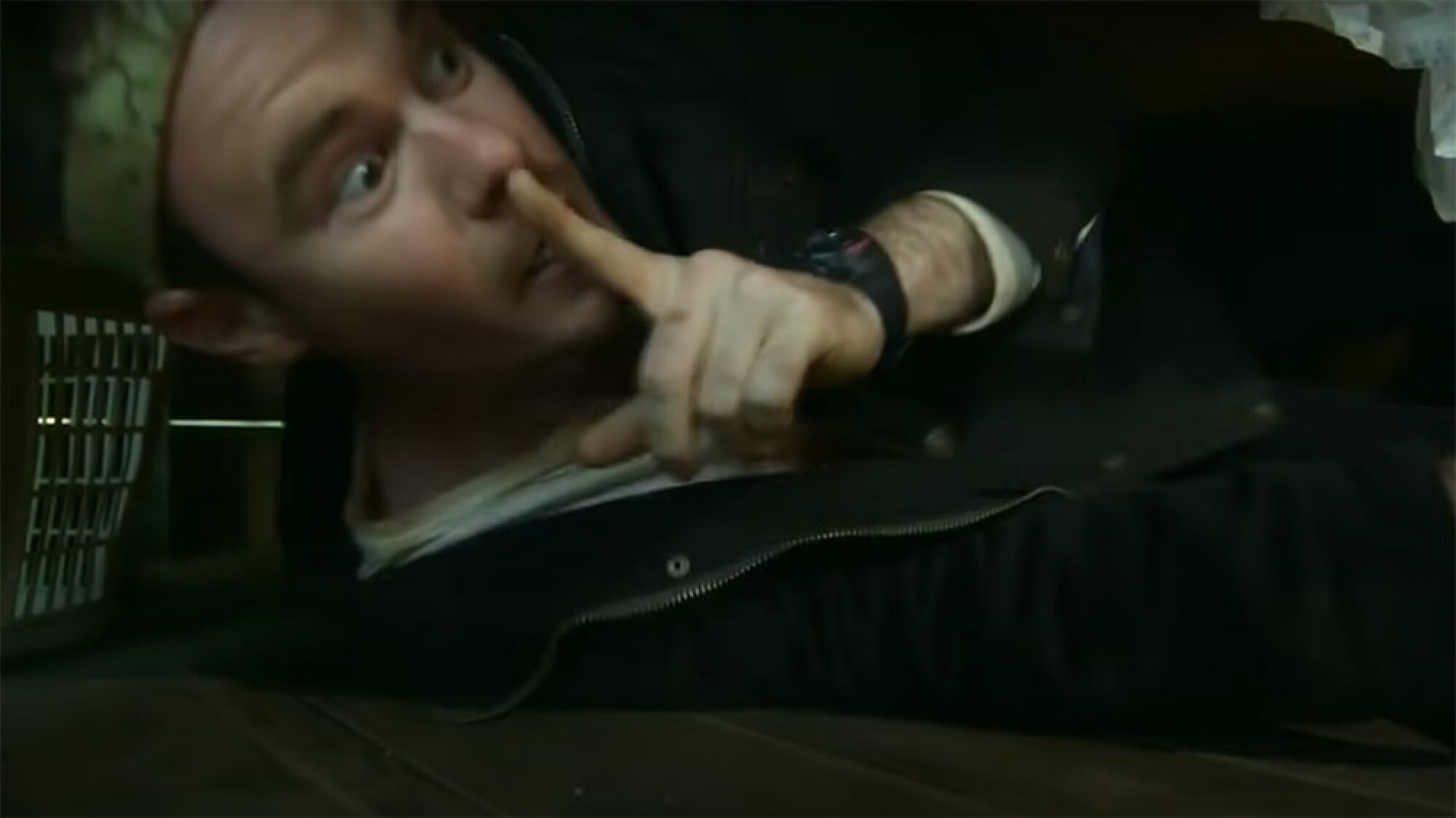 A man hiding under a bed presses his finger to his lips.
