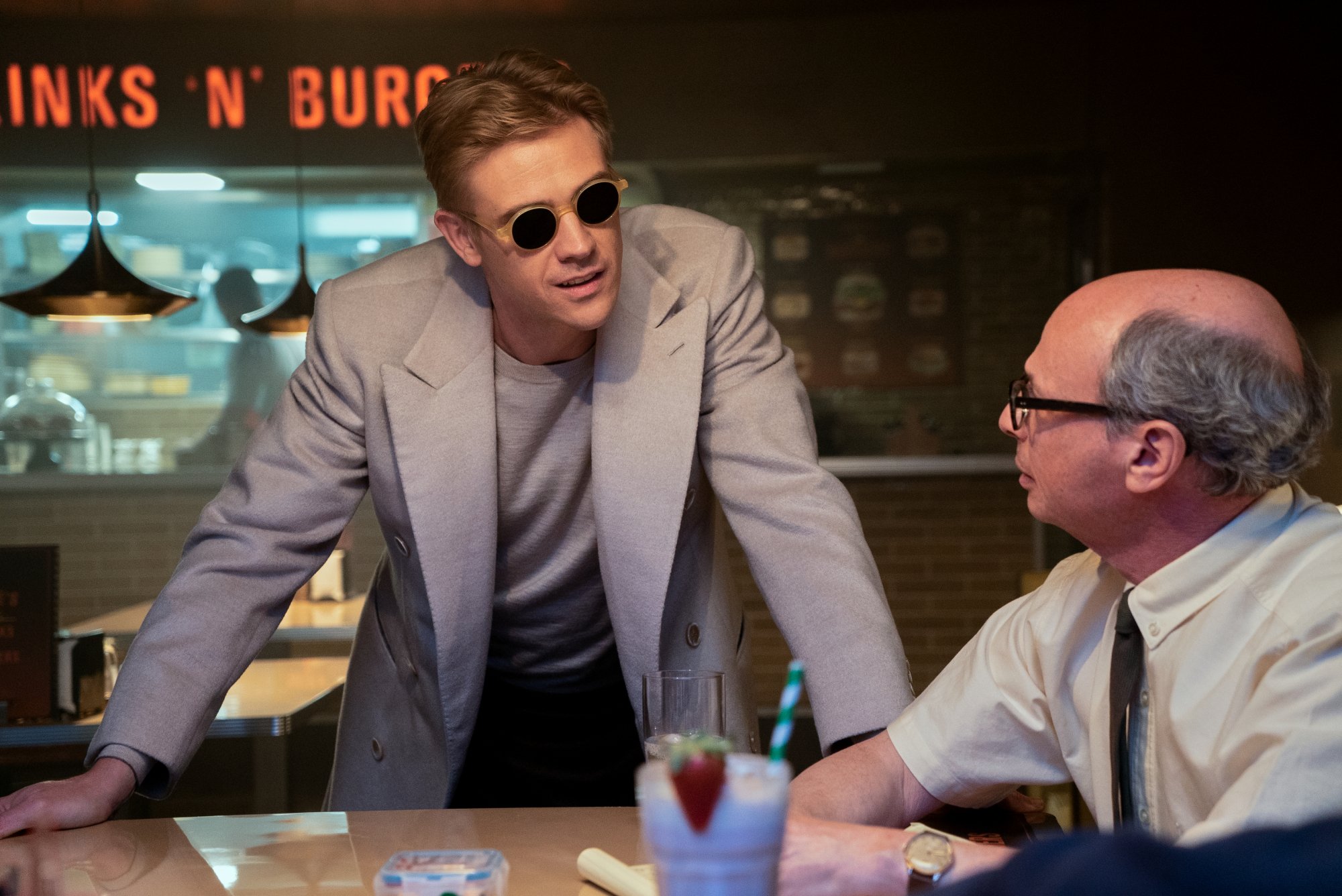 A man in sunglasses rests his hands on a diner table, talking to another man at the table.