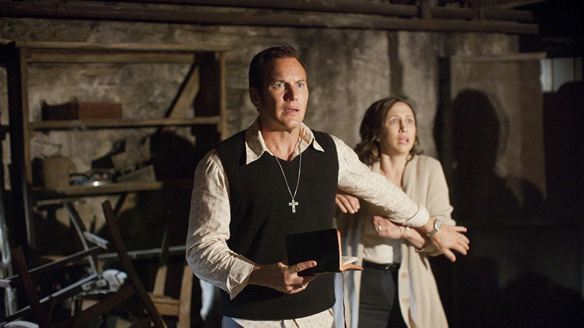 A man with a cross around his neck stands next to a woman in a basement, looking scared.