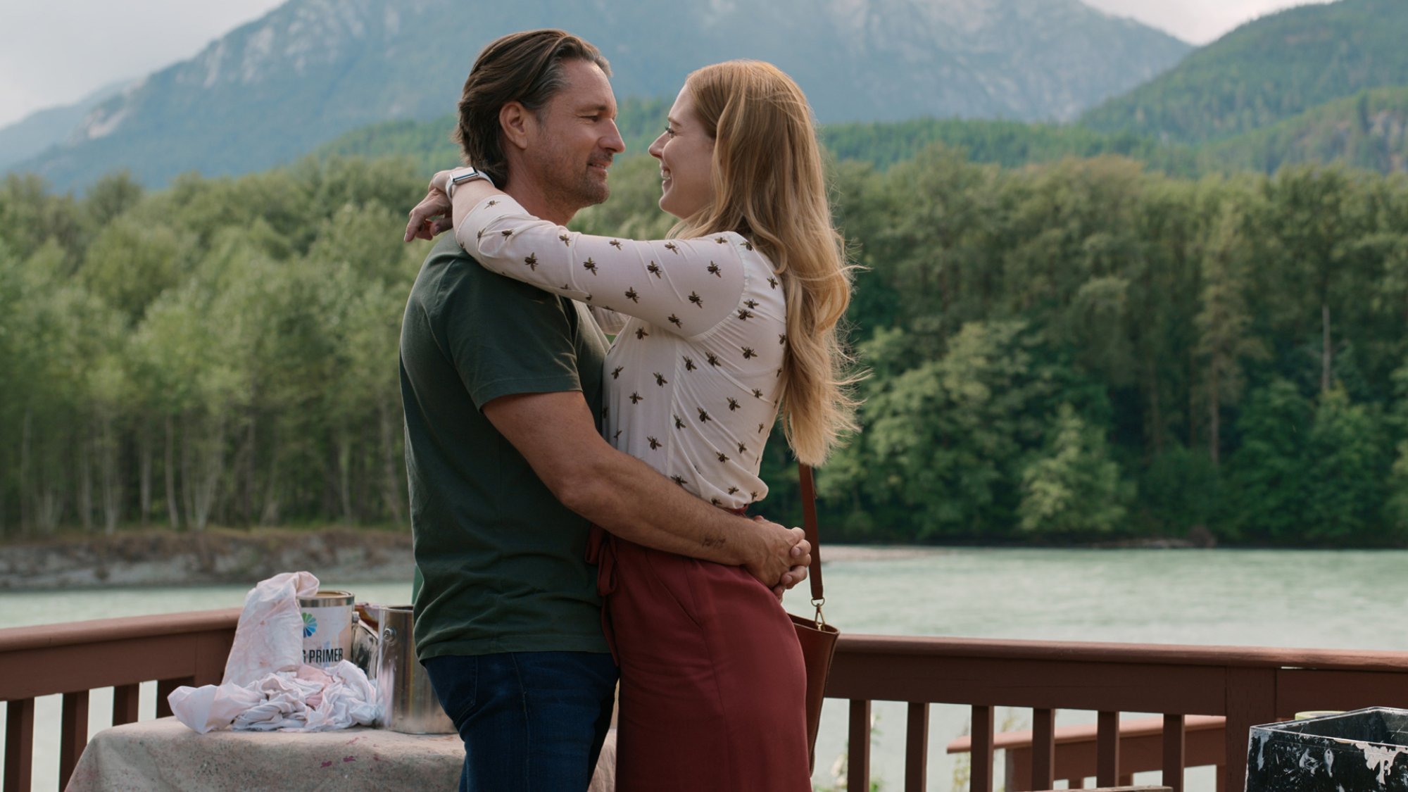 Two people embrace on a verandah beside a river.